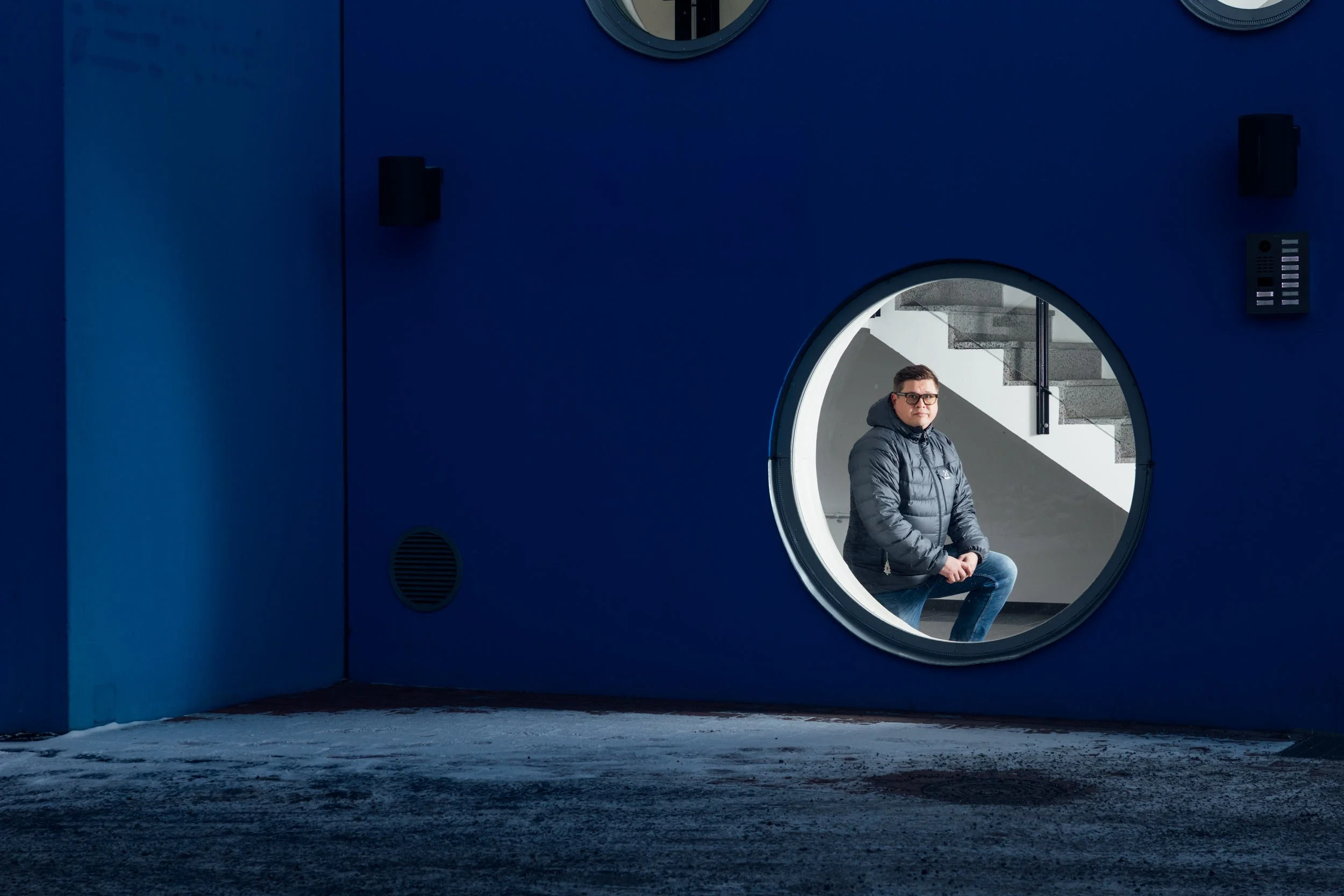 A man with glasses, wearing a gray jacket, sitting on a stool inside a circular window of a modern blue building, with stairs visible in the background.