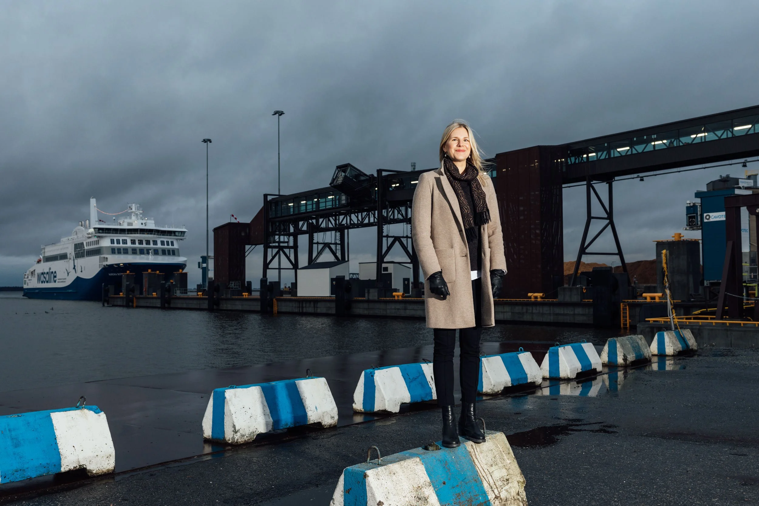 A woman standing on a concrete barrier with striped blue and white paint at a dock, with a cruise ship and industrial structures in the background, under a cloudy sky.