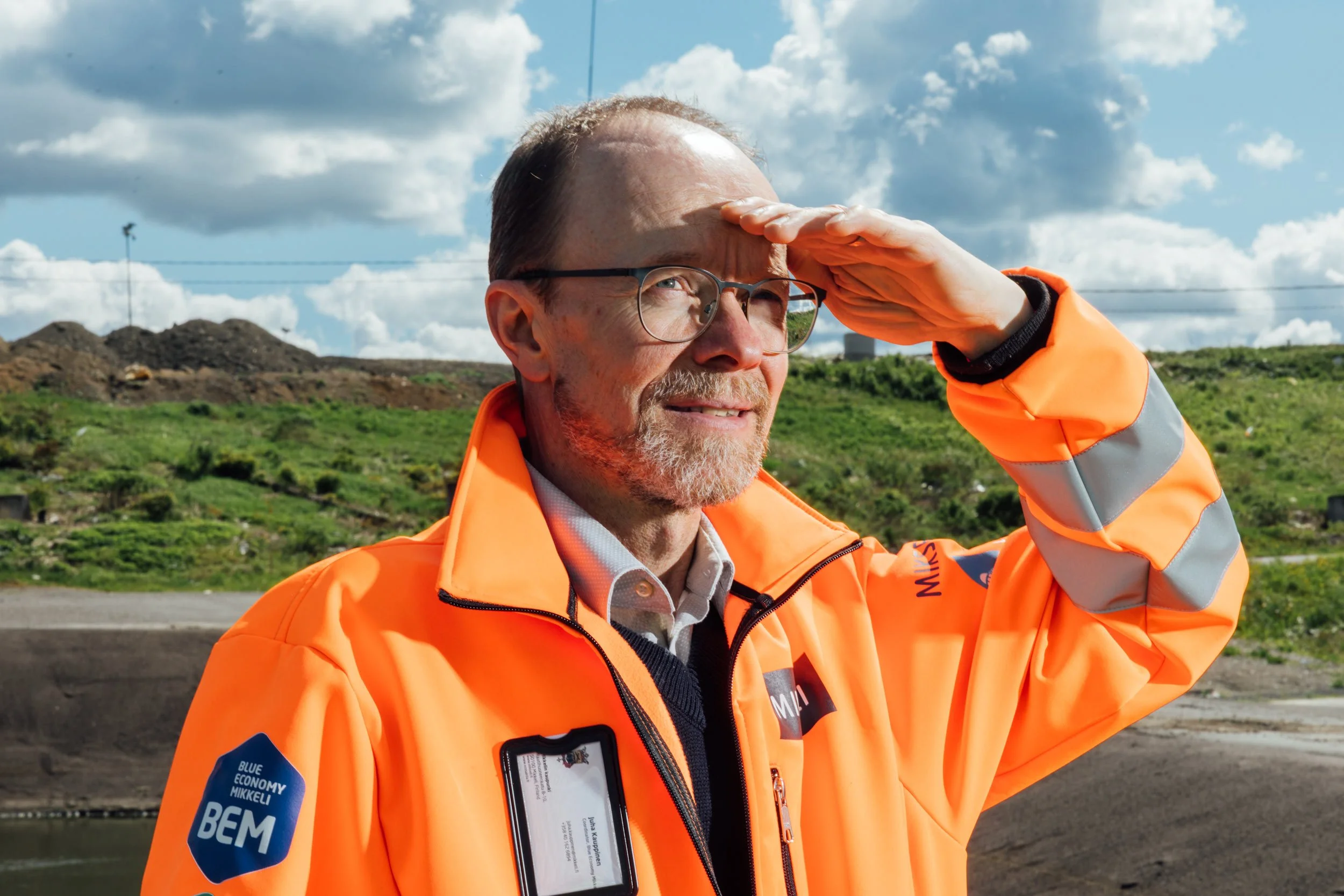 A man wearing glasses and an orange safety jacket with reflective stripes, saluting with his right hand while standing outdoors under a partly cloudy sky. The background features green hills and power lines.