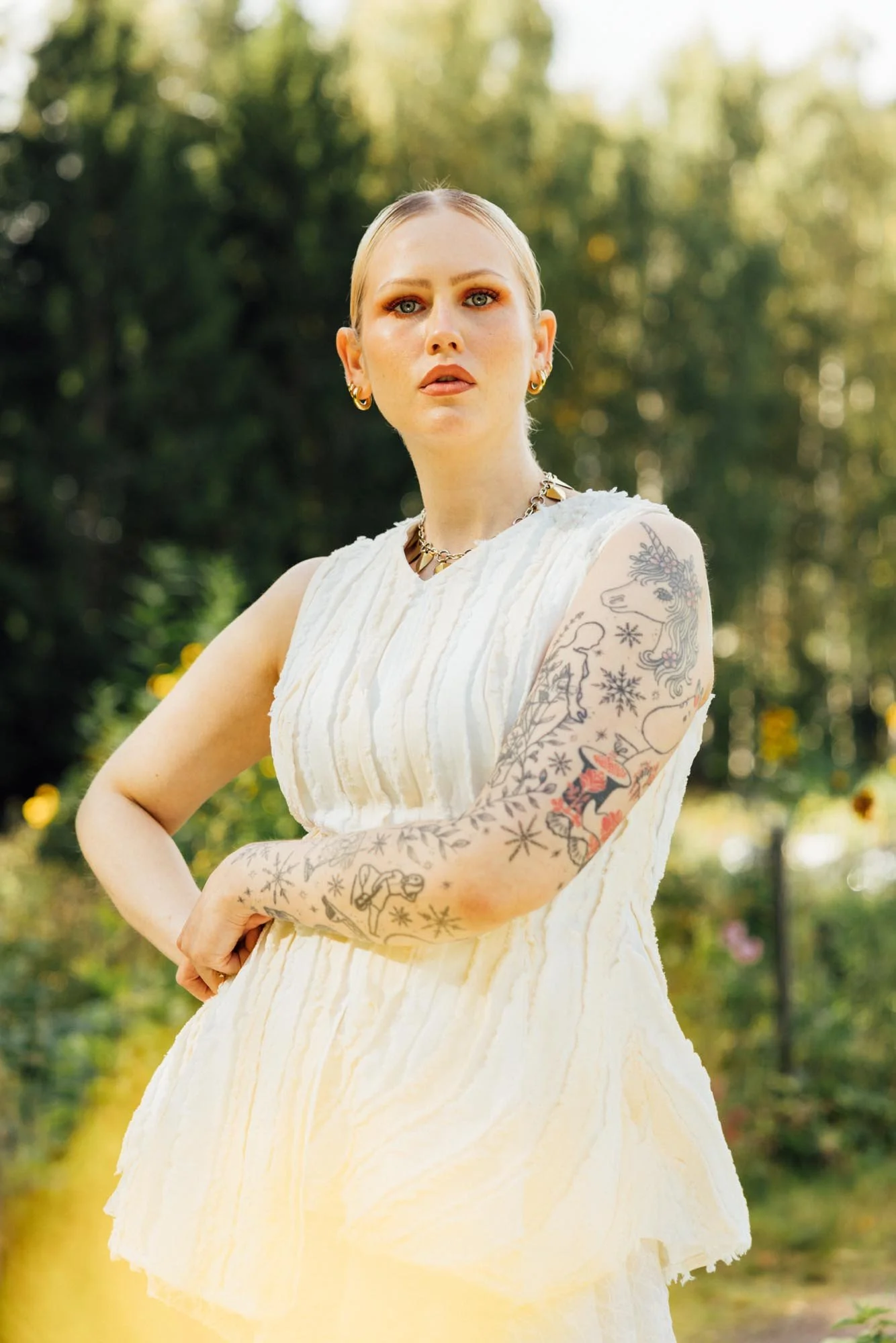 A woman with blonde hair and tattoos on her arms is standing outdoors in a yellow dress, looking into the distance with a serious expression. The background is a lush, green, blurred garden or park.