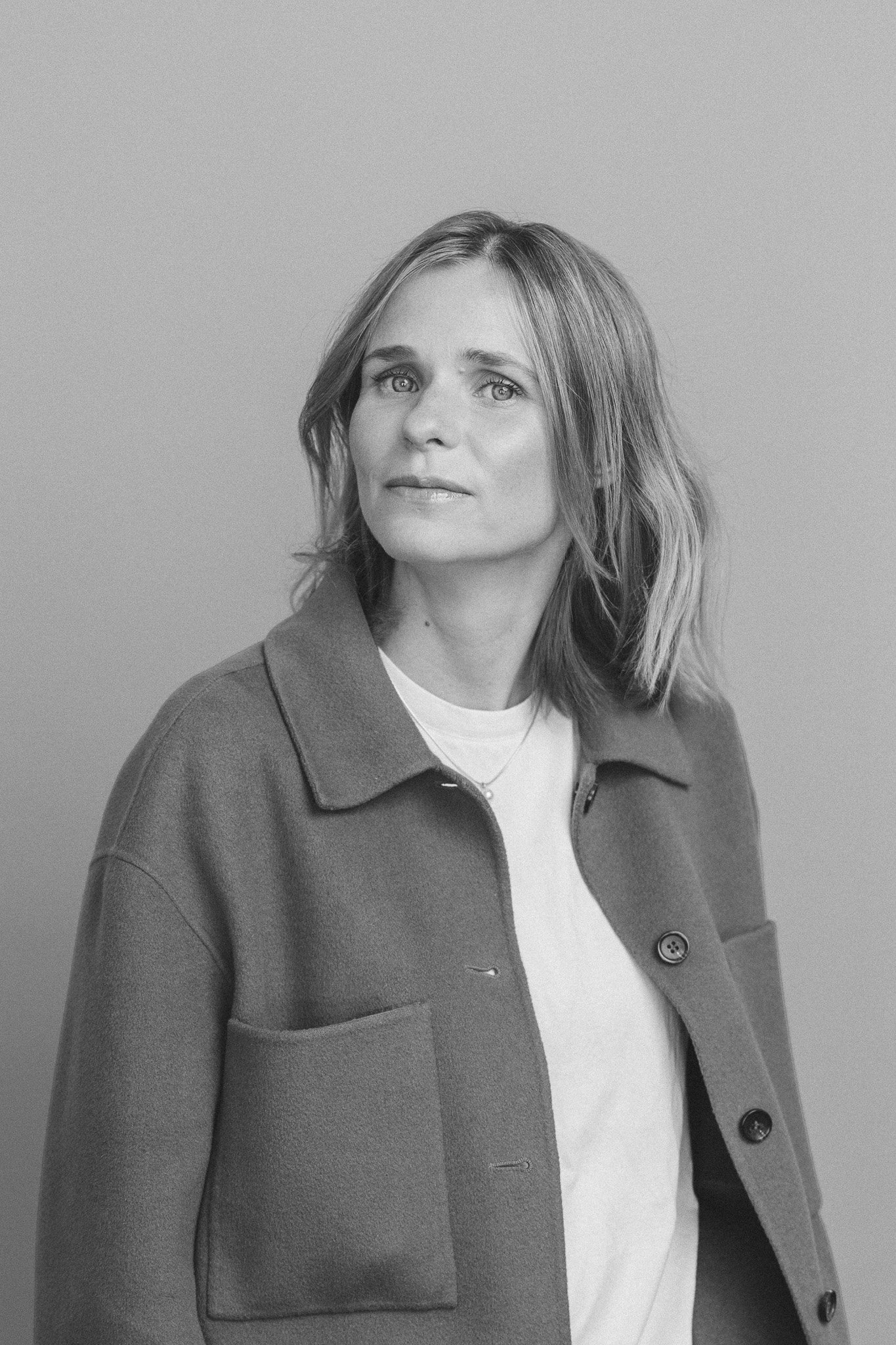 Black and white portrait of a woman with shoulder-length hair wearing a coat over a sweater, looking slightly off-camera.