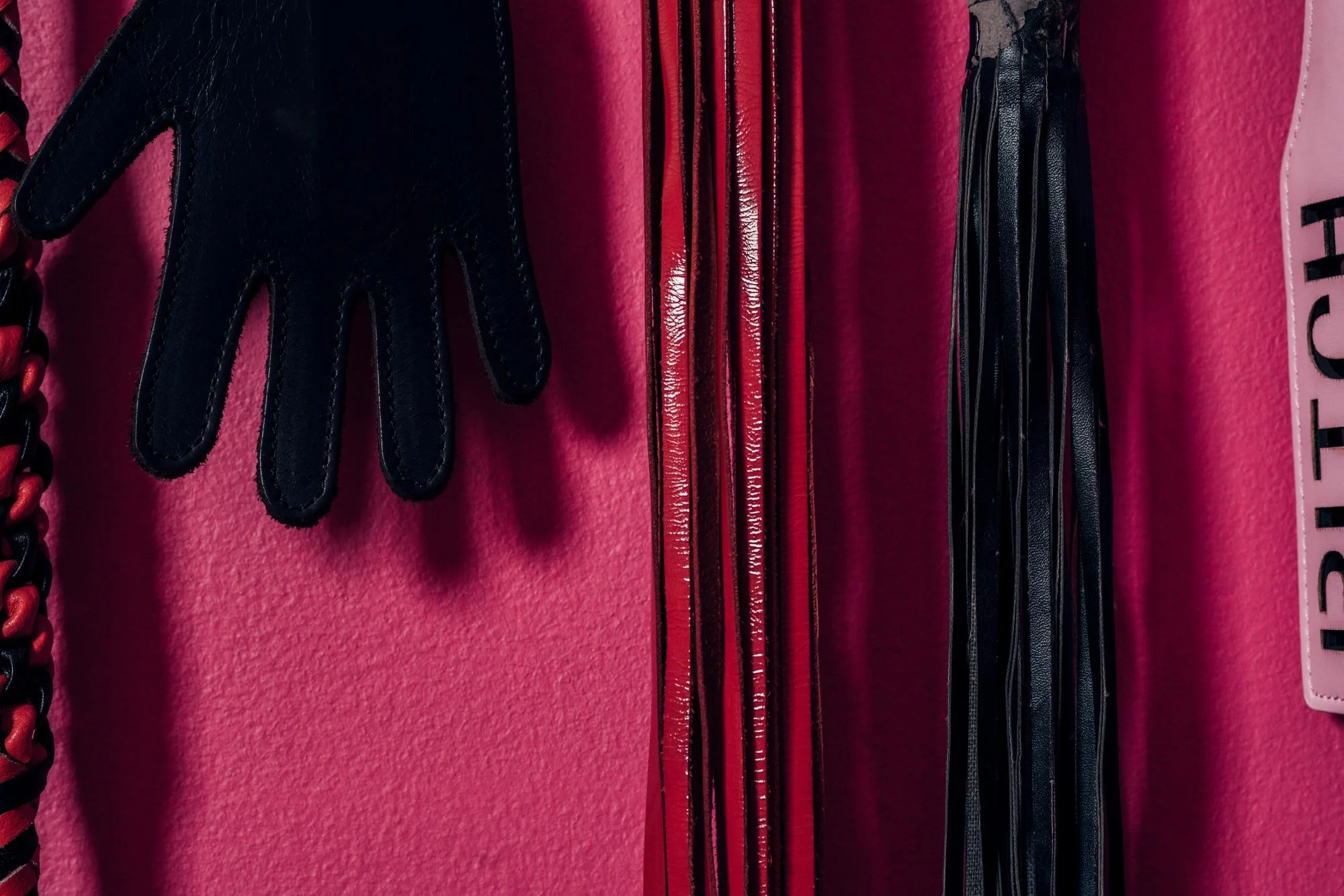 Close-up of a pink wall with various fashion accessories hanging, including a black leather glove, red and black chain strap, and black and red zippers.