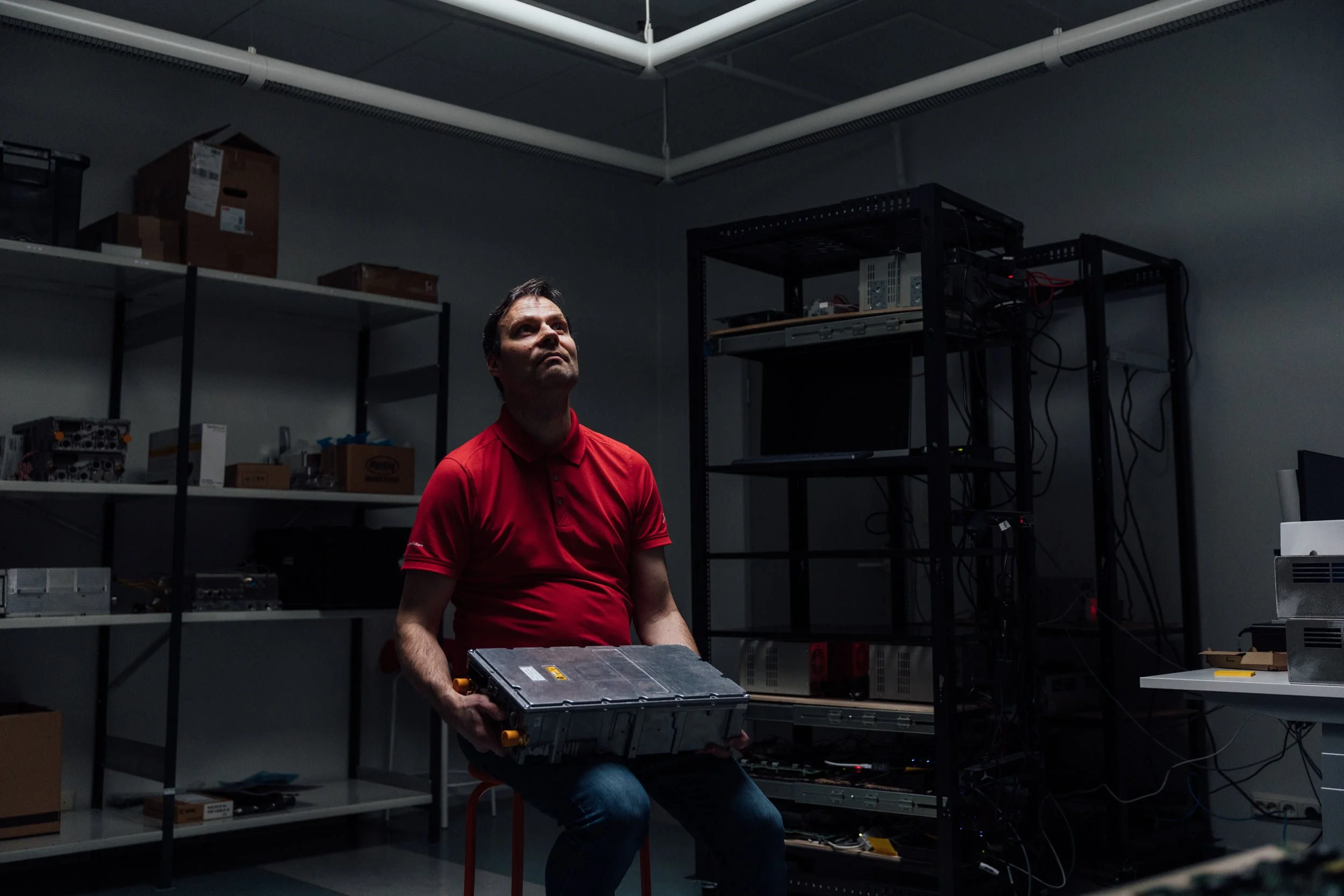 A man in a red polo shirt sitting on a stool in a dark, cluttered room holding a large electronic device.