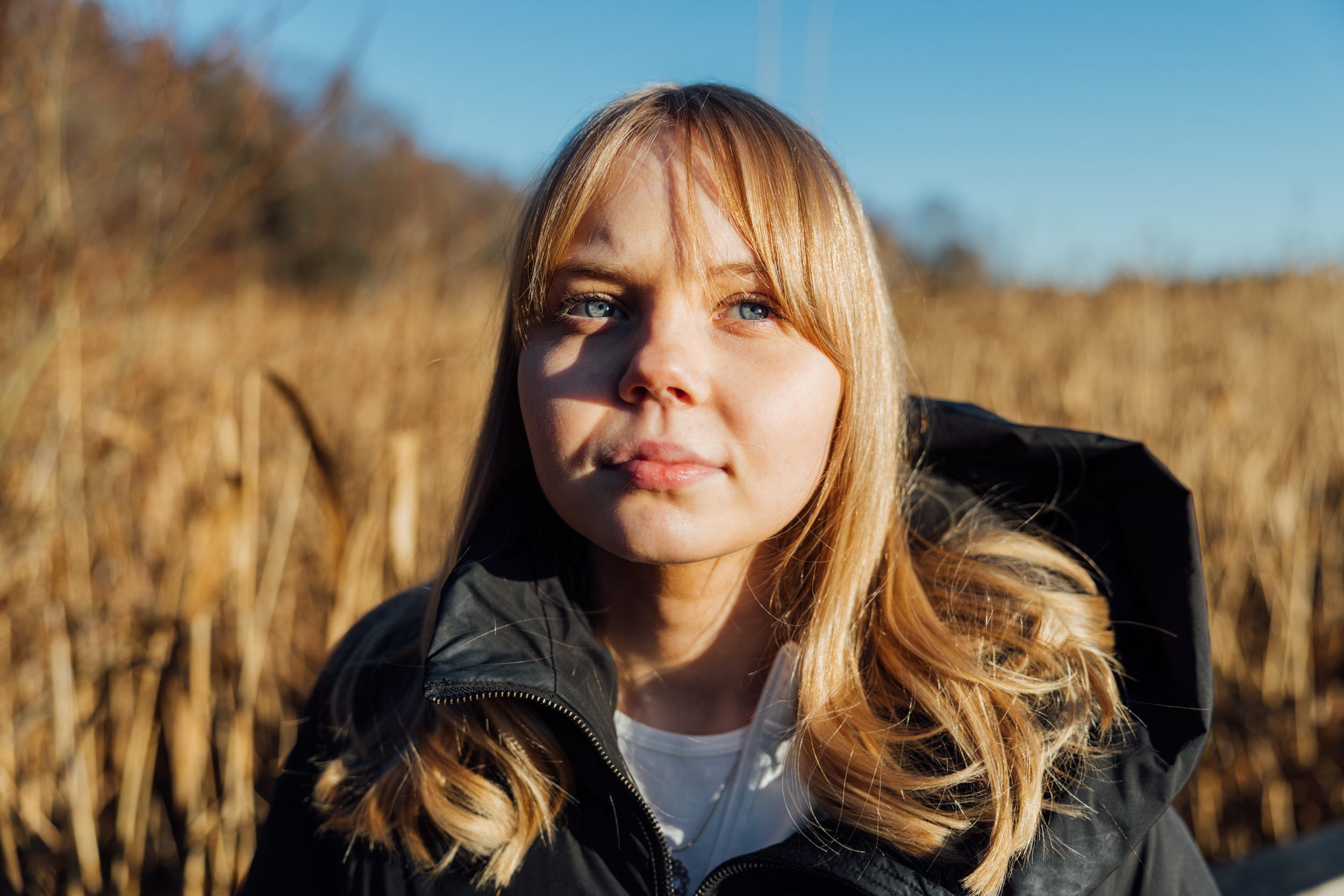 Close-up of a young woman with long, wavy, strawberry blonde hair and blue eyes, outdoors with a blurred field in the background, wearing a black jacket.
