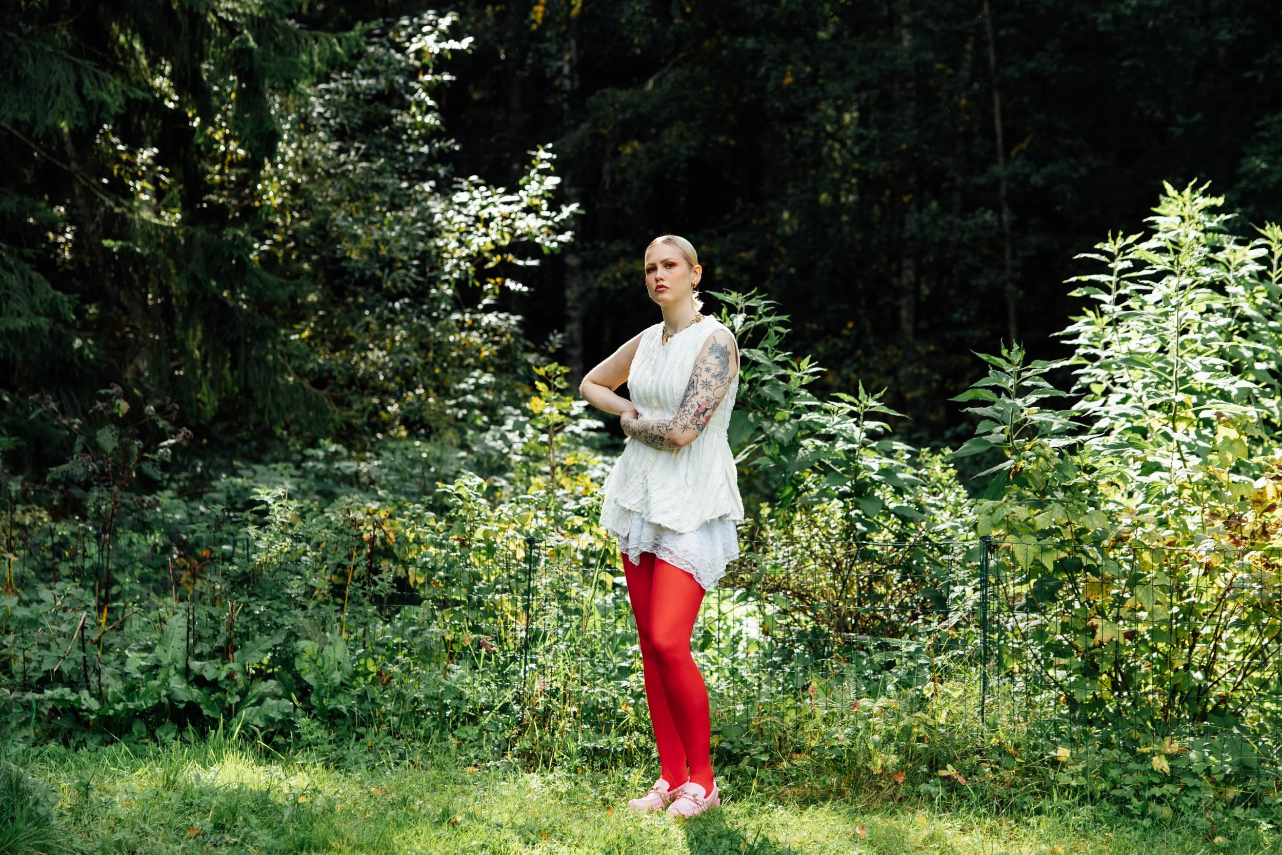 Young woman with blonde hair and tattoos on her arms standing outdoors in a lush green forest, wearing a white dress and bright red tights.
