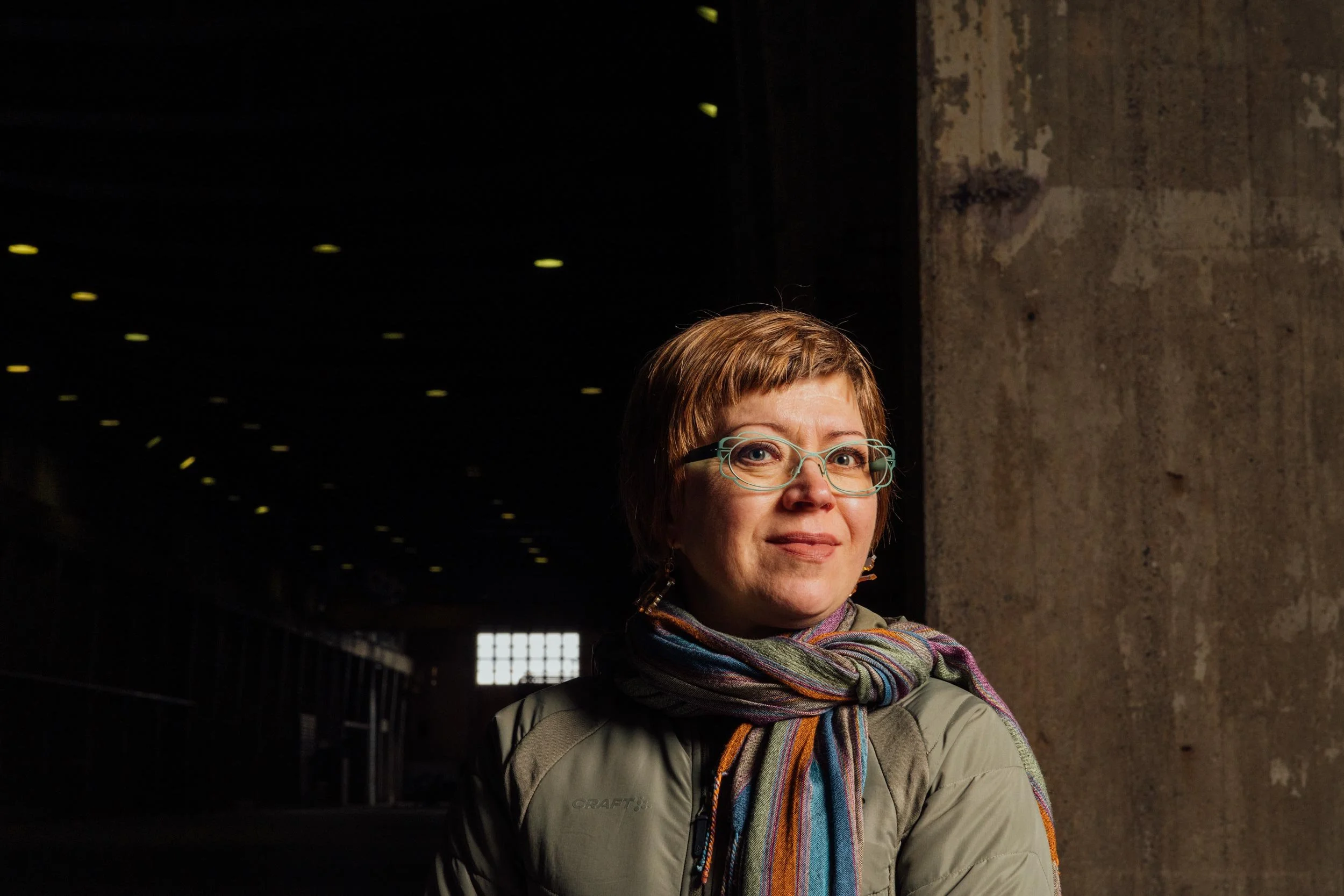 A woman with short brown hair and glasses, wearing a tan jacket and a colorful scarf, standing outdoors near a concrete wall with dark sky and small yellow lights in the background.