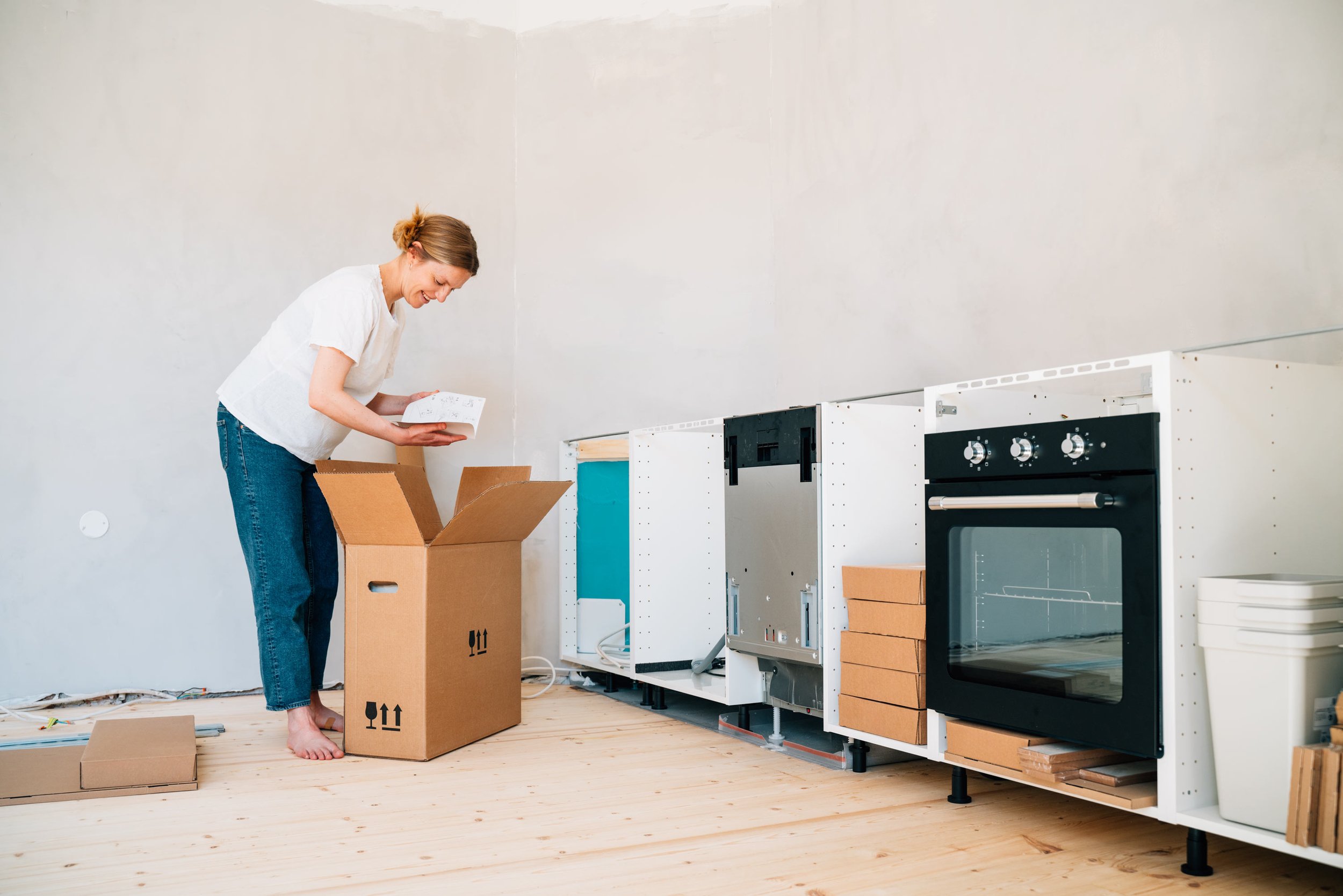 A woman with light brown hair tied in a bun, wearing a white shirt and jeans, is smiling and looking at papers while standing next to a cardboard box. She is barefoot. The room has unfinished light wood flooring and plain white walls. There are partially assembled kitchen cabinets and oven on the right side of the image. Some cardboard boxes and packaging materials are on the floor.