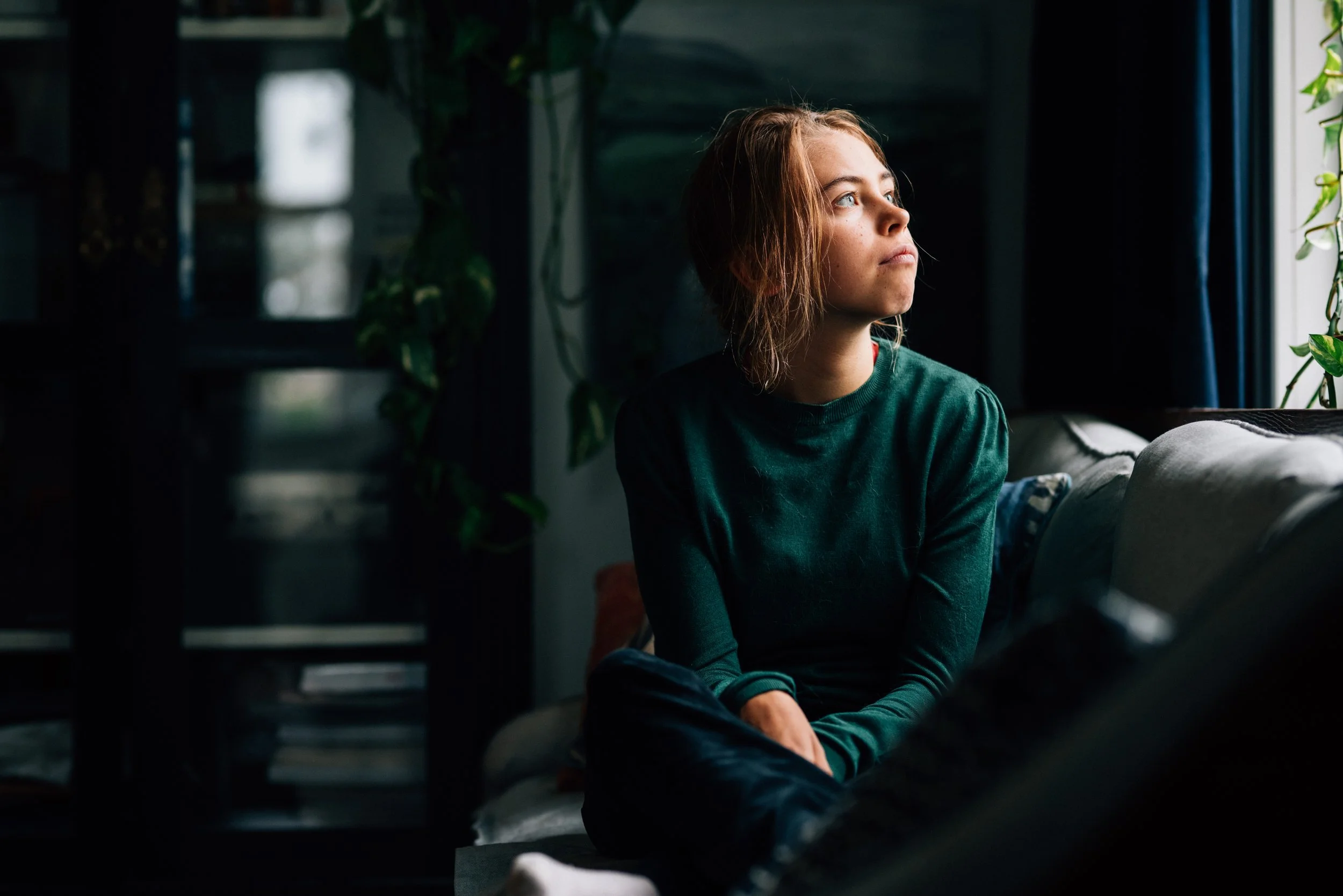 A young woman with light brown hair looking out a window, sitting indoors with sunlight illuminating her face.