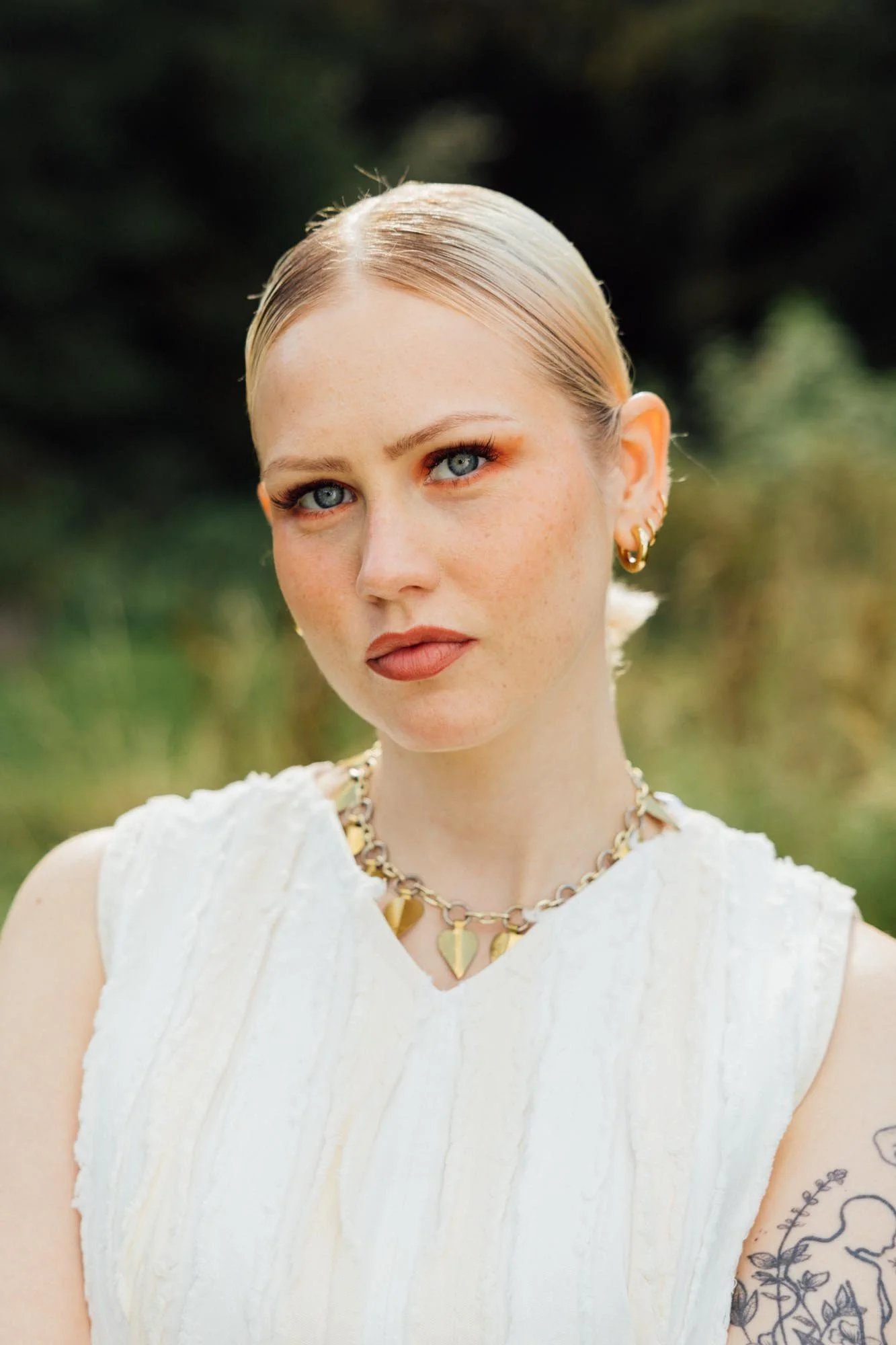 A woman with blonde hair styled back, blue eyes, wearing gold jewelry including a necklace and earrings, dressed in a white sleeveless top, standing outdoors with greenery in the background.