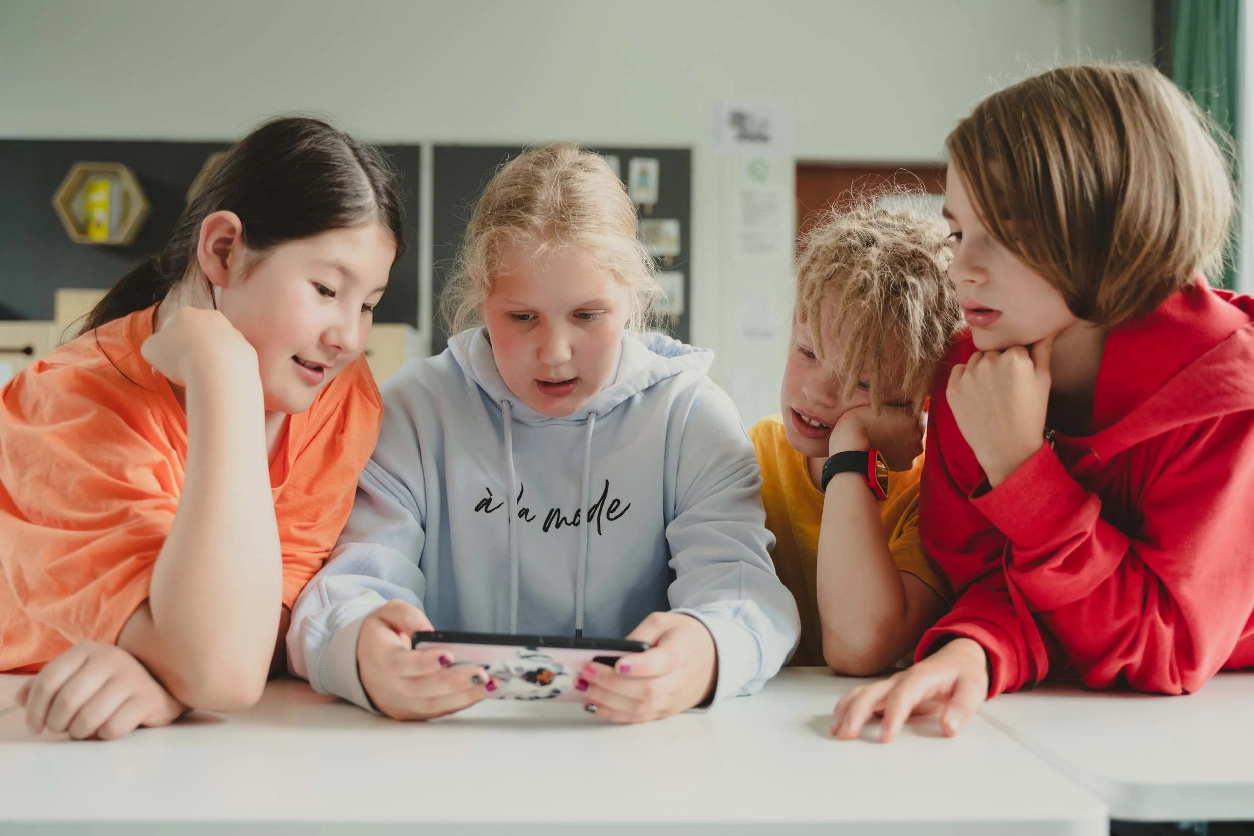 Group of five children gathered around a table, looking at a phone together, in a classroom setting.