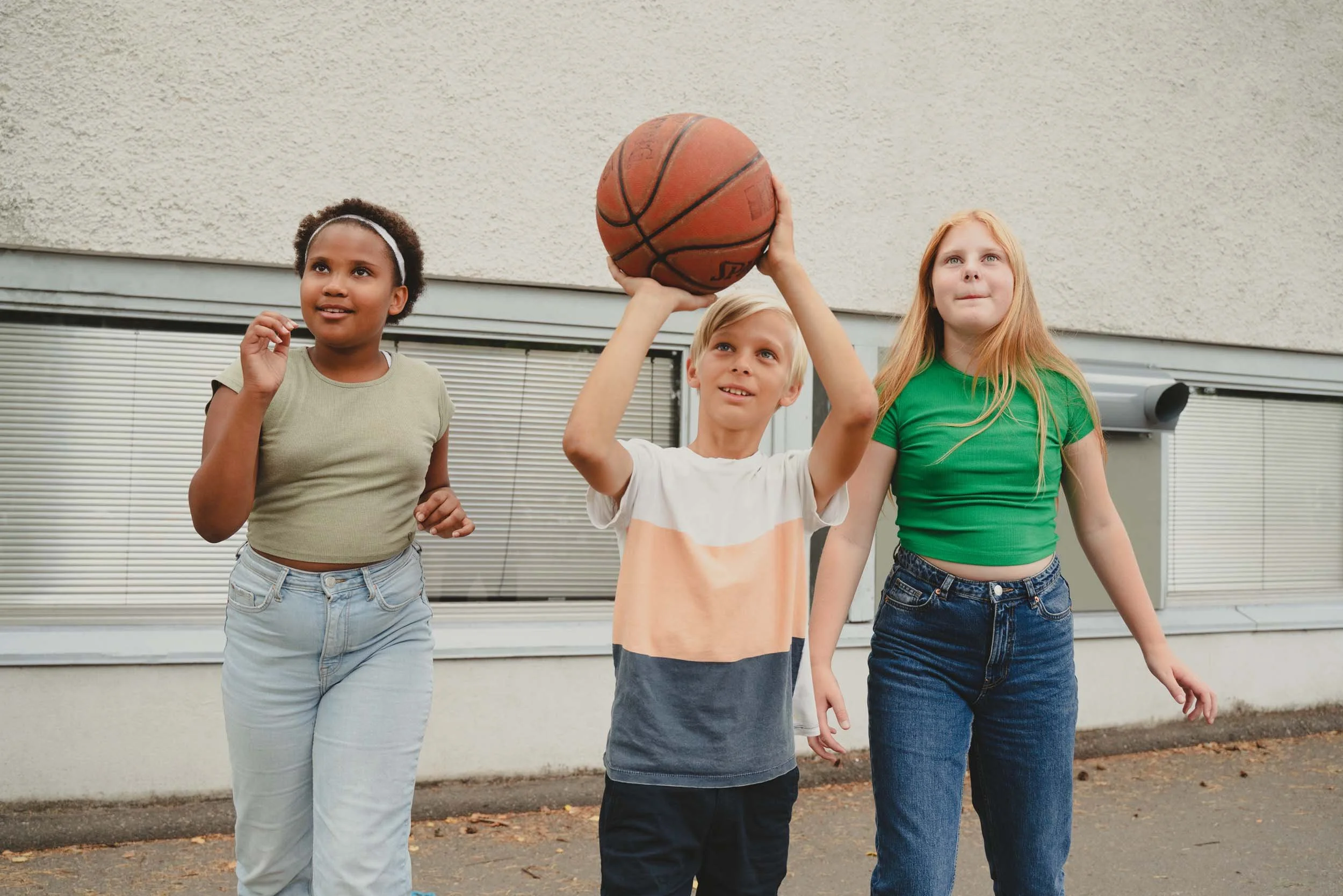 Three children outdoors, one boy holding a basketball, two girls standing next to him, all casually dressed.