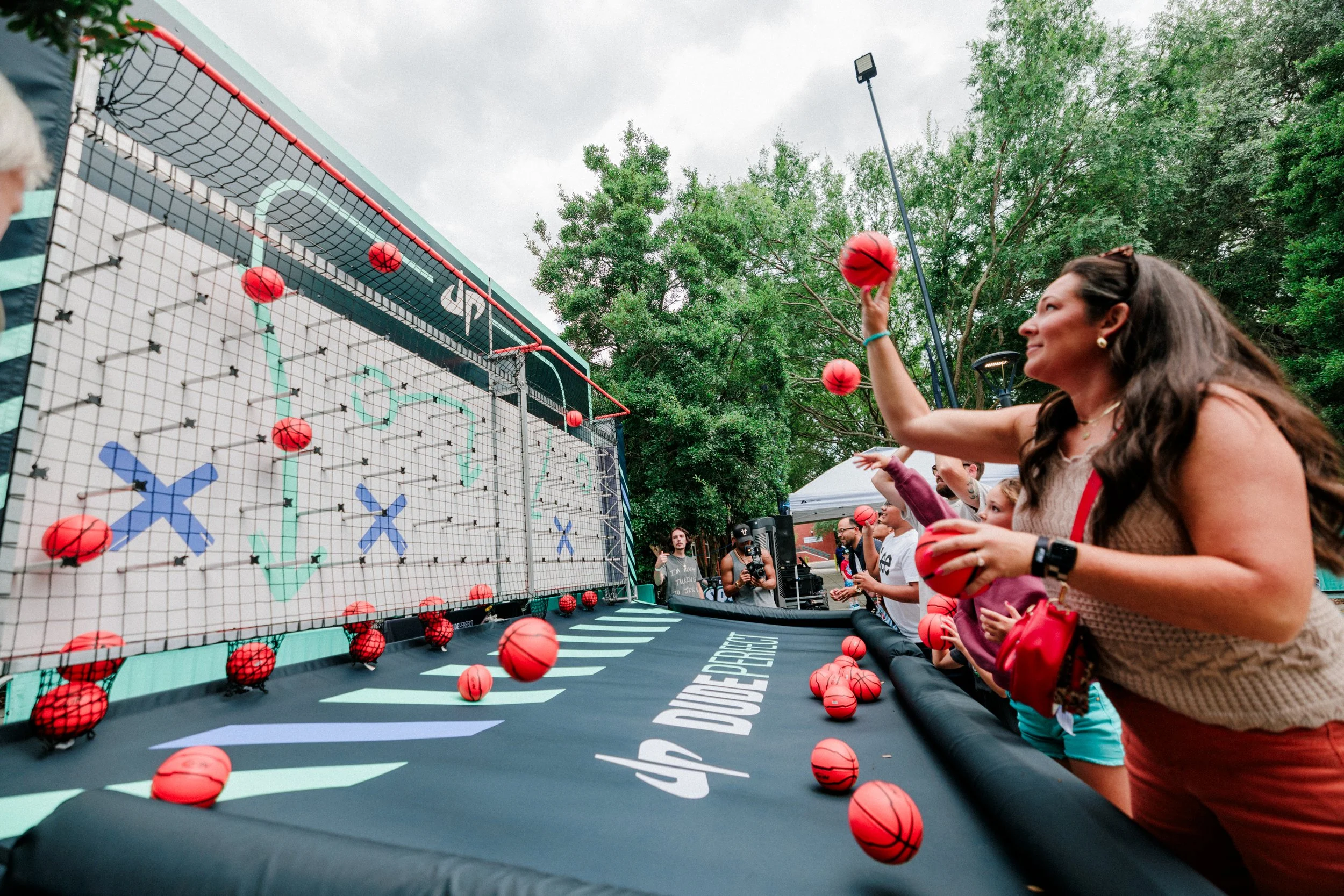 People participating in a basketball shooting game at an outdoor event, aiming at a large target with a net. The game involves throwing small red basketballs at the target.
