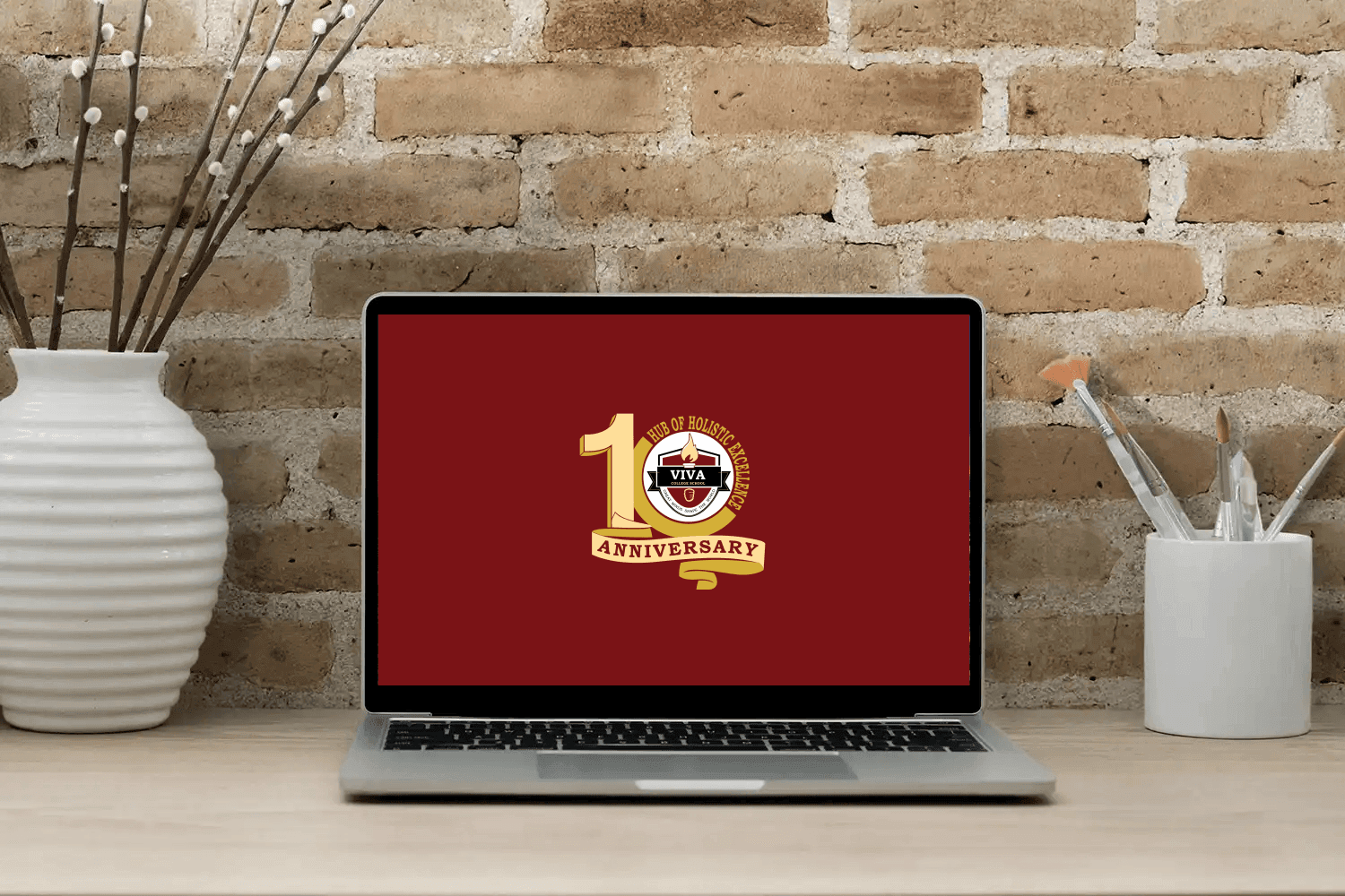 Laptop on a wooden desk displaying a 10th anniversary logo for VIVA College School with a brick wall background, flanked by white vases holding decorative sticks and paintbrushes.