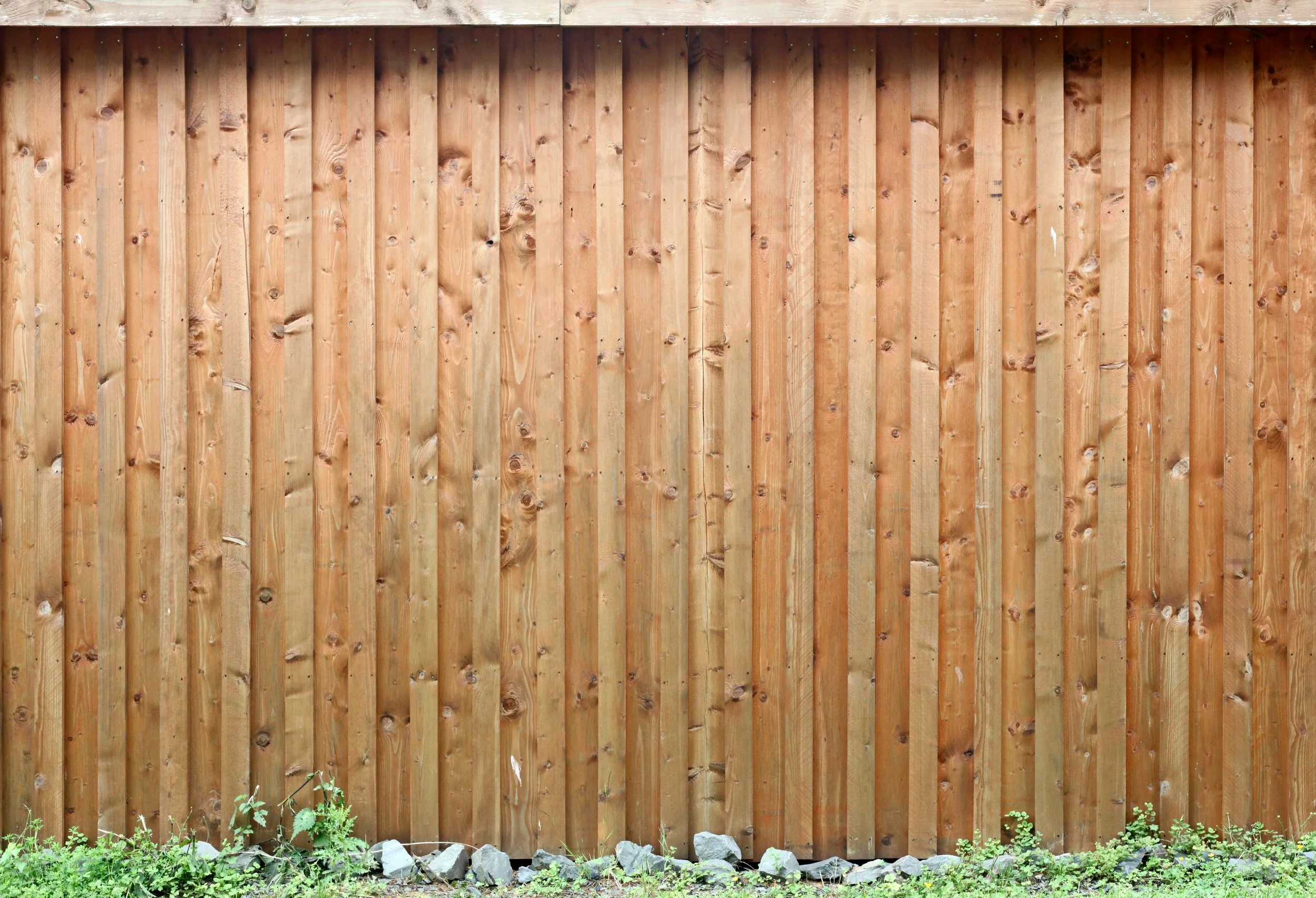 A wooden privacy fence with vertical planks, with rocks and green grass at the base.