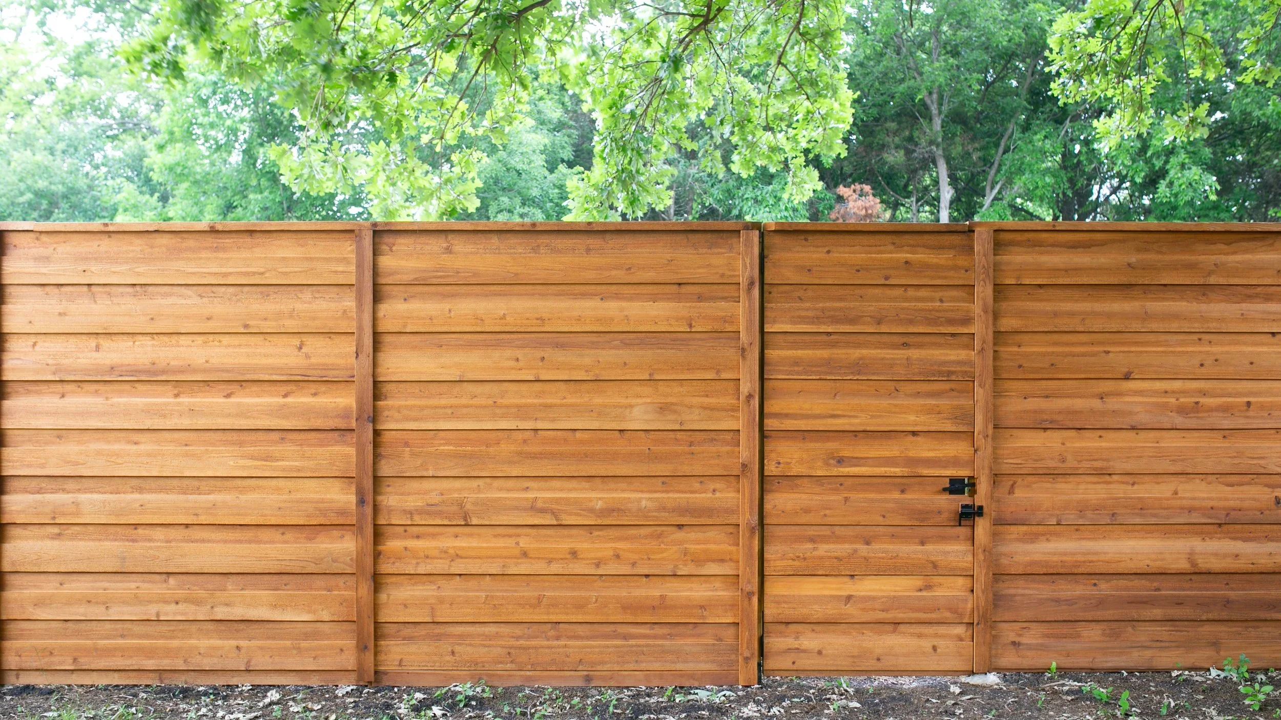 A wooden fence with horizontal slats and vertical support beams, with a small gate locked with black latches, set against a backdrop of green trees and foliage.