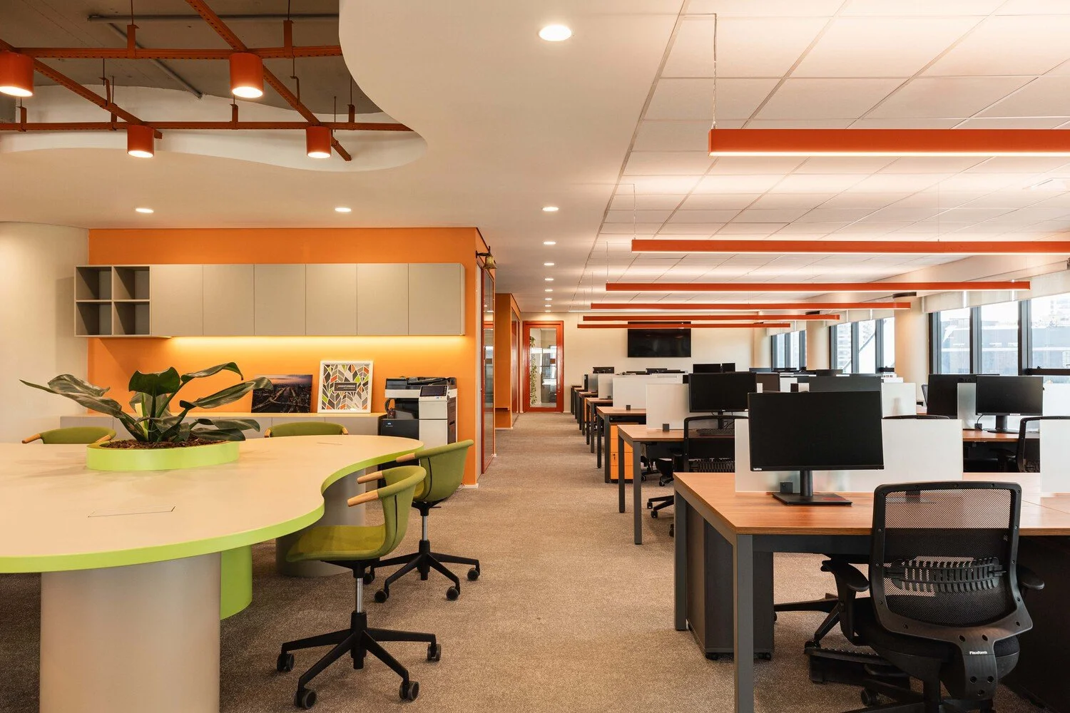 Modern open-plan office with rows of desks, computer monitors, and large windows, featuring orange accents and a casual seating area with a green plant.