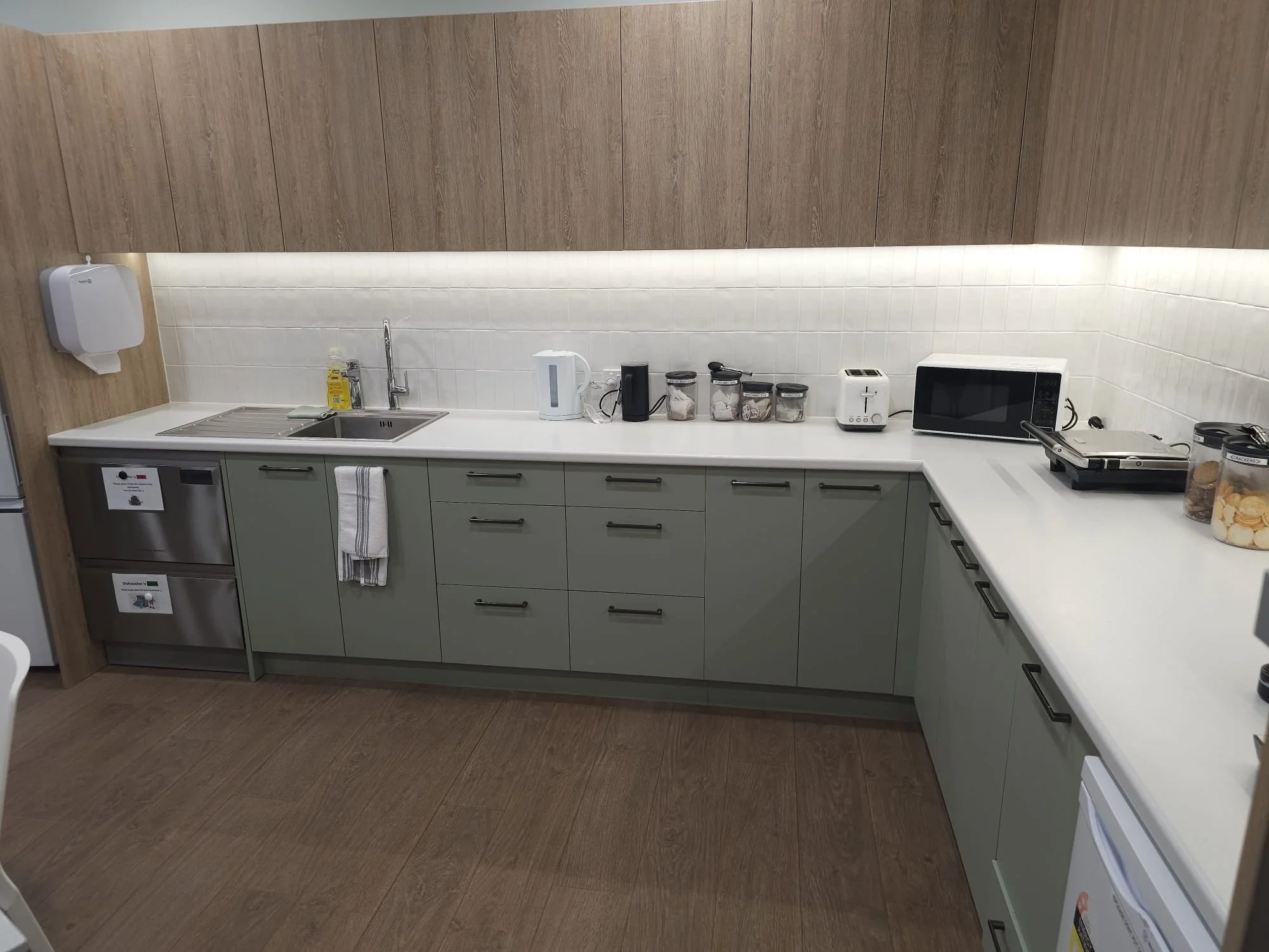 Modern kitchen with light green lower cabinets, white countertop, and white tiled backsplash. Contains sink, electric kettle, toaster, microwave, sandwich press, and jars of snacks.