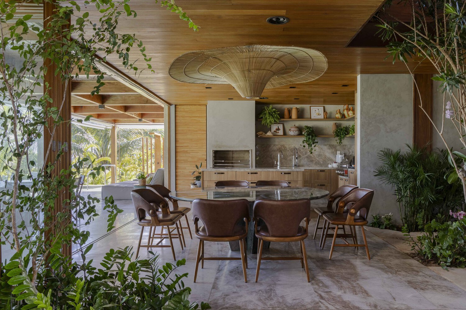 Indoor dining area with a round marble table, six brown leather chairs, and a built-in outdoor kitchen with greenery and palm trees visible outside.