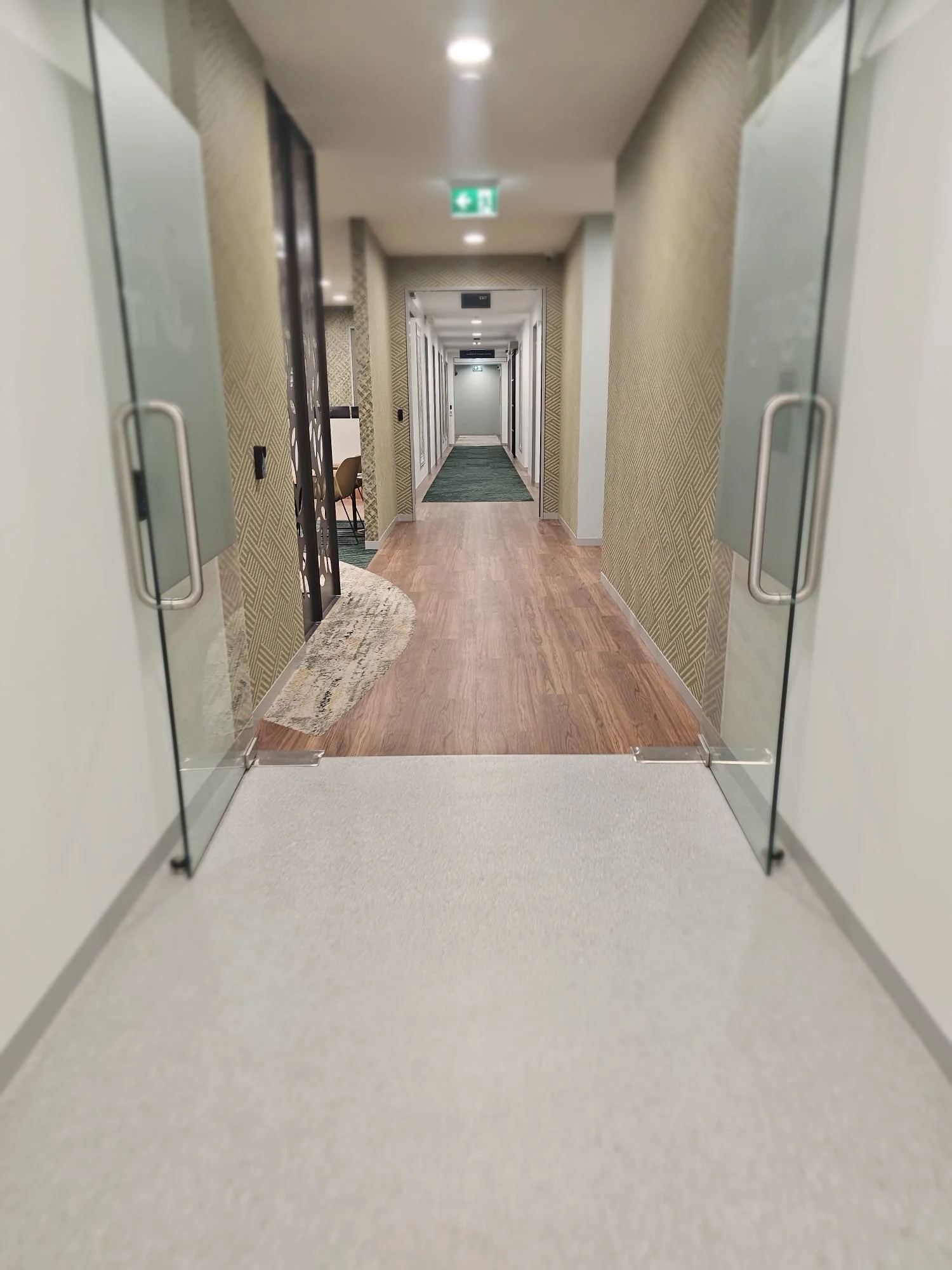 View of a hotel corridor seen through glass doors, with patterned wallpaper, wooden flooring, and a green exit sign overhead.