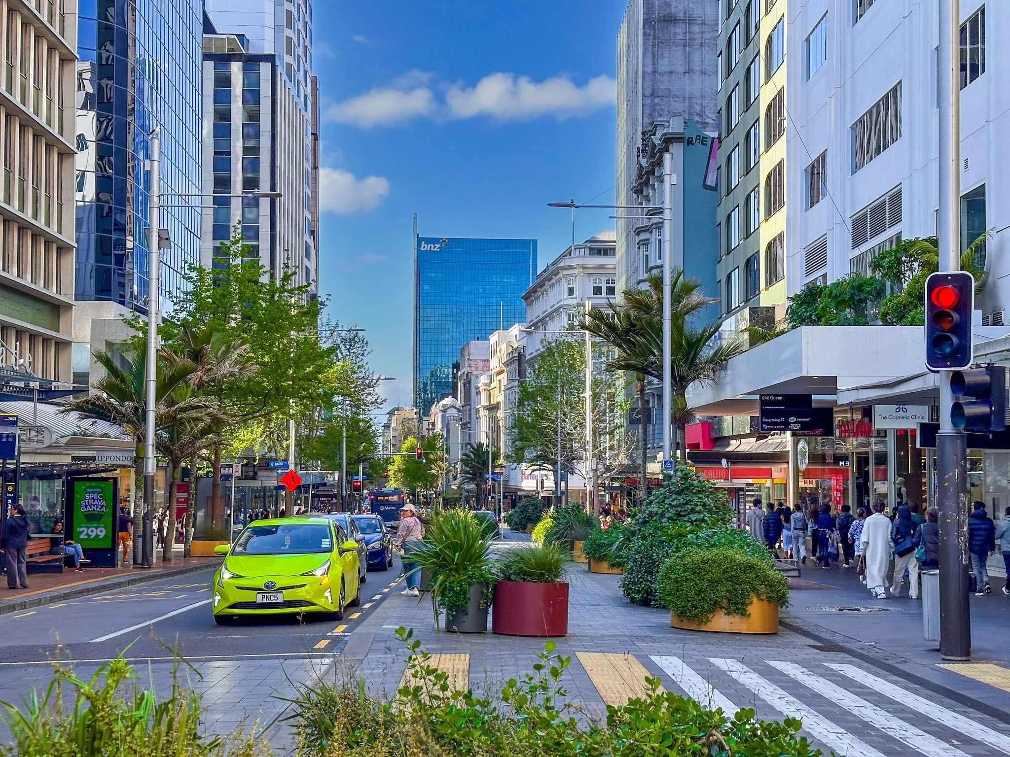 A busy city street with tall buildings, green trees, and large planters lining the sidewalk. There are cars, including a bright green one, and pedestrians walking on the sidewalk. Traffic lights and street signs are visible, and a clear blue sky is above.