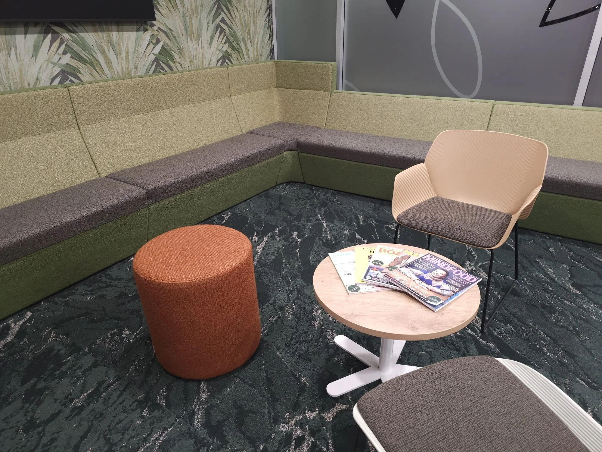 A modern waiting area with an L-shaped green and gray sofa, an orange ottoman, a beige armchair with gray cushion, a small round wooden table with magazines, and a patterned dark green carpet.