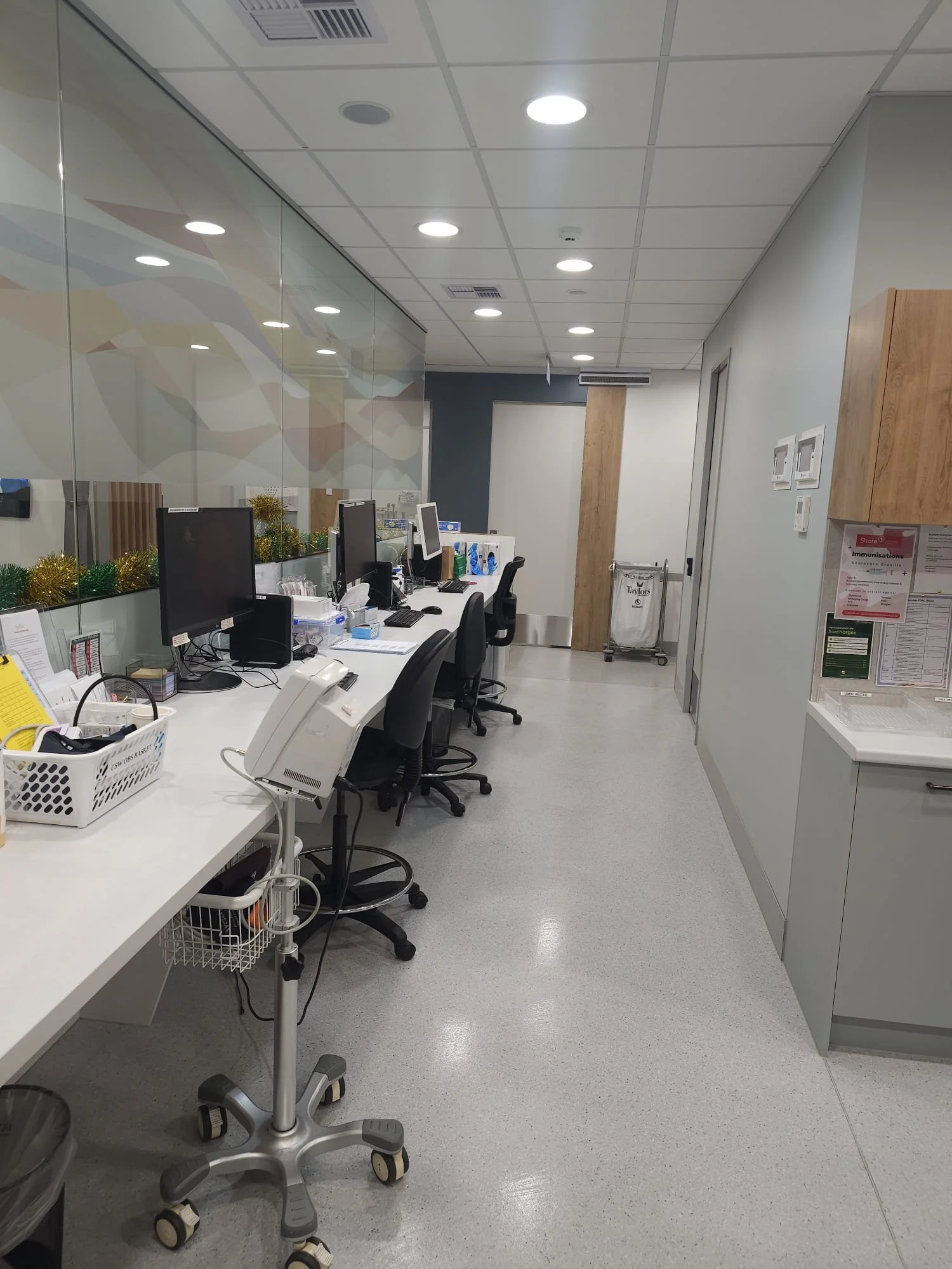 A hospital reception area with a long white counter, computer monitors, telephones, and office chairs. The area is decorated with holiday tinsel near the window, and there are medical notices on the wall.