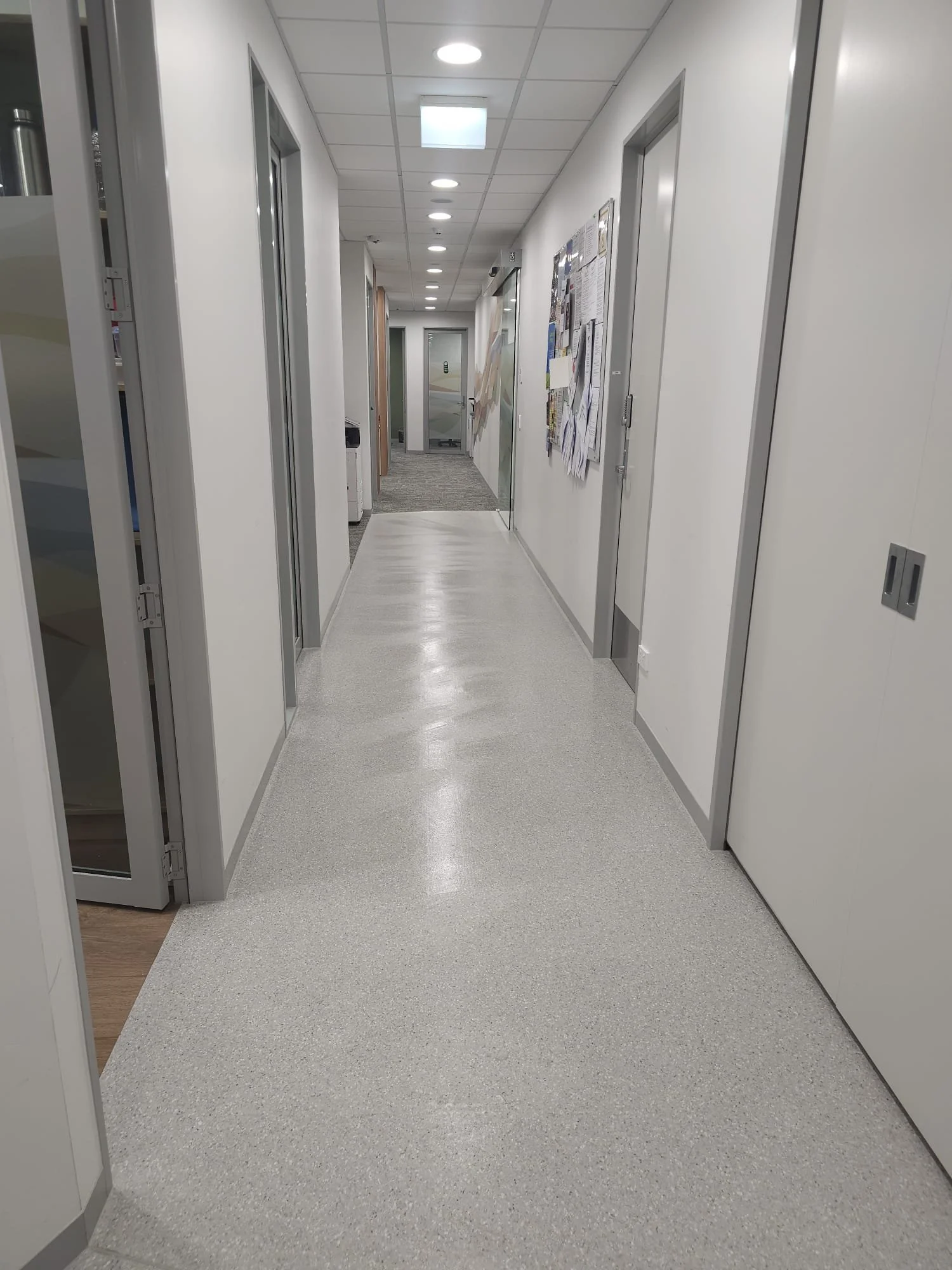 Empty office hallway with closed and open doors on both sides, bulletin boards with papers and flyers on the right wall, ceiling lights, and a door at the end of the hallway.