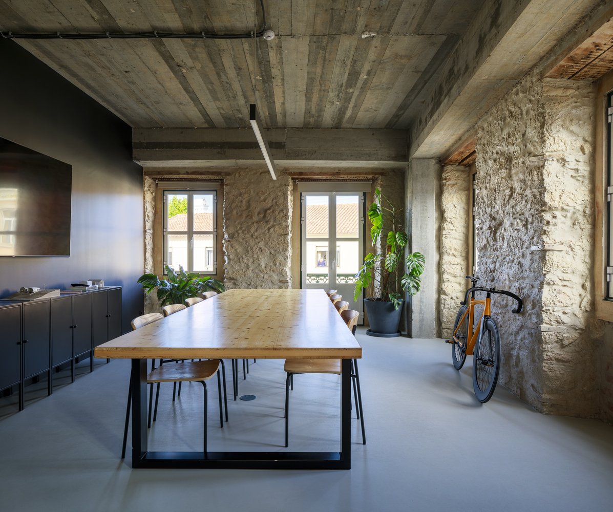 Modern meeting room with a large wooden table, chairs, potted plants, and a bicycle, featuring exposed stone and concrete walls and ceiling, with windows letting in natural light.