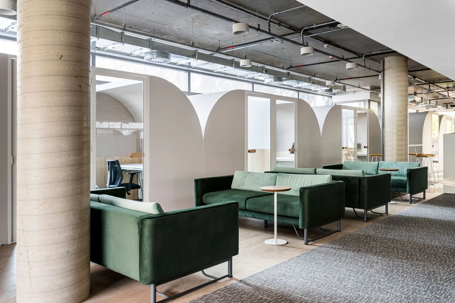 Modern lounge area with green velvet sofas, small side tables, and semi-private study booths in the background with white walls and wooden handles in a contemporary office space.