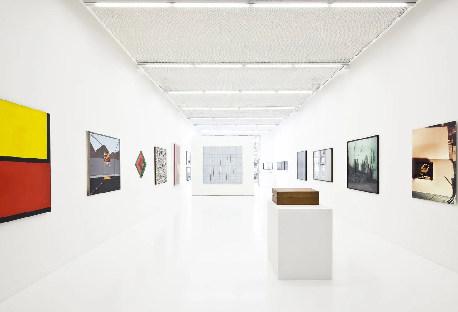 A modern art gallery with white walls, featuring various colorful and abstract paintings and framed artwork, with a white pedestal and a wooden bench in the center.