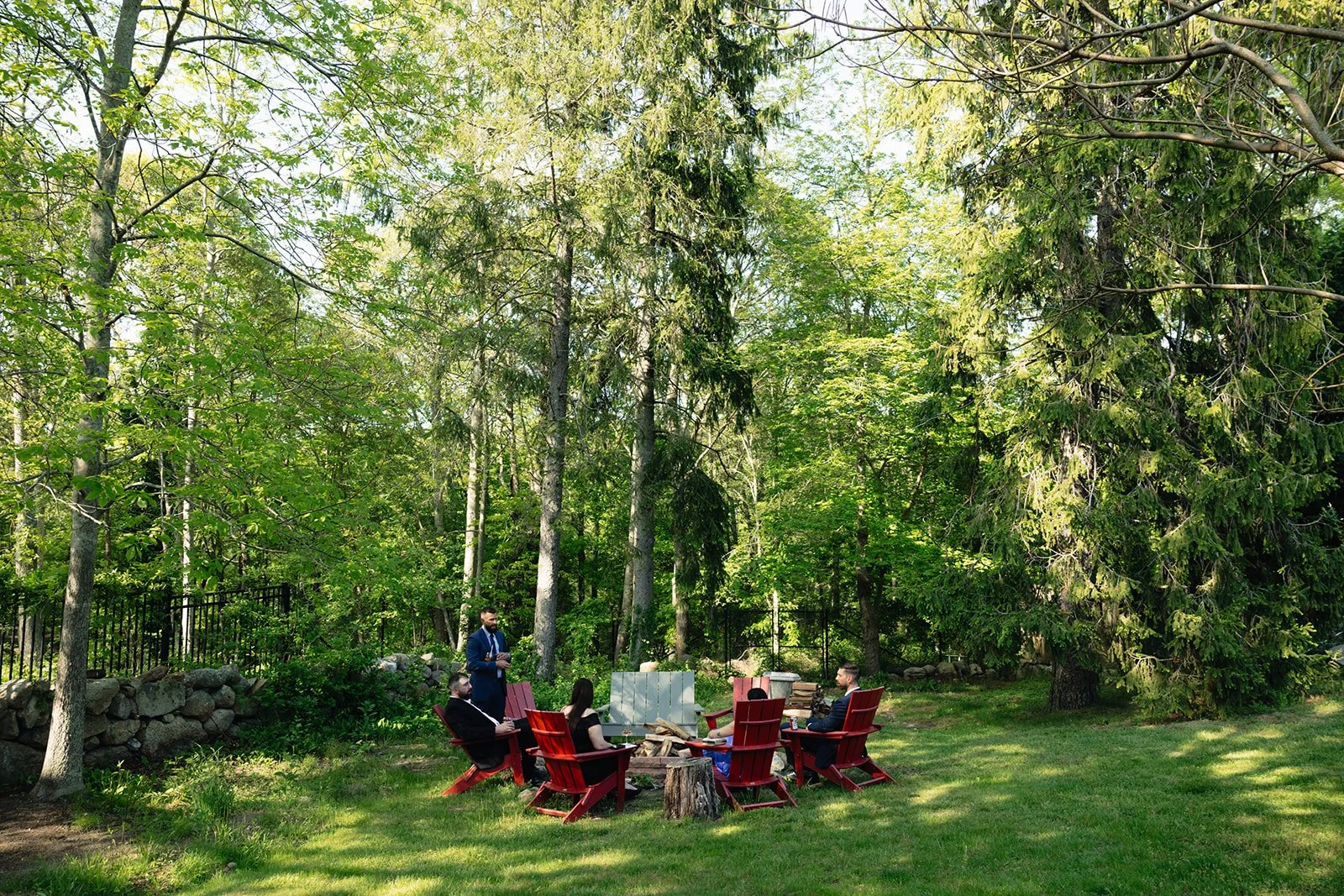 People gathered around a firepit in a green outdoor setting, surrounded by tall trees and a grassy lawn during a wedding at Lambert's Cove Inn on Martha's Vineyard.