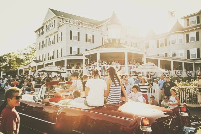 A large crowd gathered outside a historic white hotel decorated with patriotic bunting for a parade or celebration. People are sitting and standing around a vintage convertible car parked on the street. The scene appears to be during a sunny day.