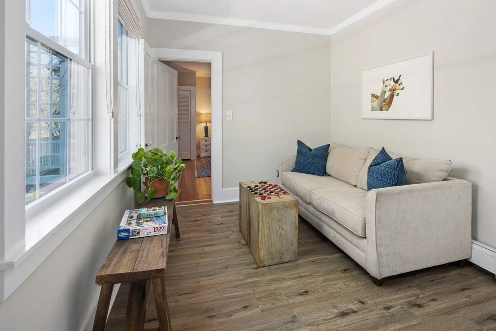 A cozy living room with a beige sofa, blue pillows, a small wooden coffee table, and a wooden side table with a plant, a board game, and magazines. There are three windows with curtains and a door leading to a hallway with a lamp and a side table.