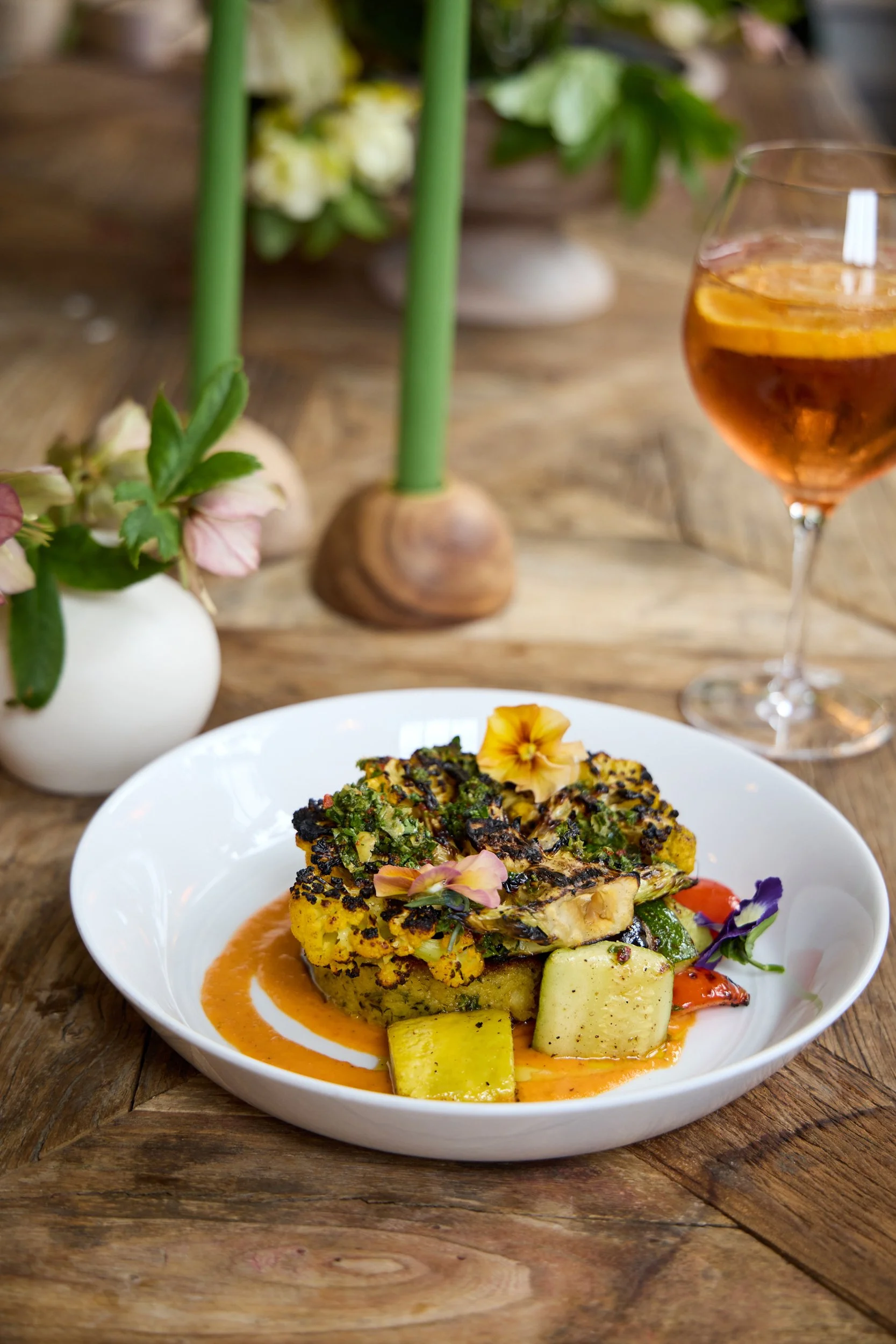 A plated dish consisting of a baked casserole or frittata topped with herbs and edible flowers, served with roasted potatoes, on a white plate. In the background, there is a glass of pink-colored beverage with lemon slices, a vase with flowers, and a wooden table.