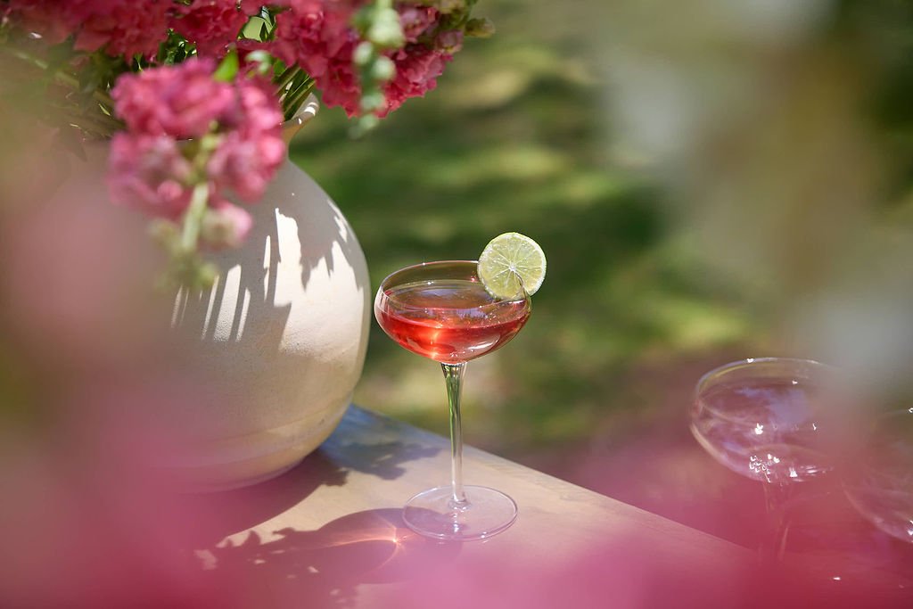 A pink cocktail with a lime slice garnish in a coupe glass on a wooden surface, with a large white vase filled with pink flowers in the background, outdoors with blurred greenery at Mermaid Bar at Lambert's Cove Inn on Martha's Vineyard.