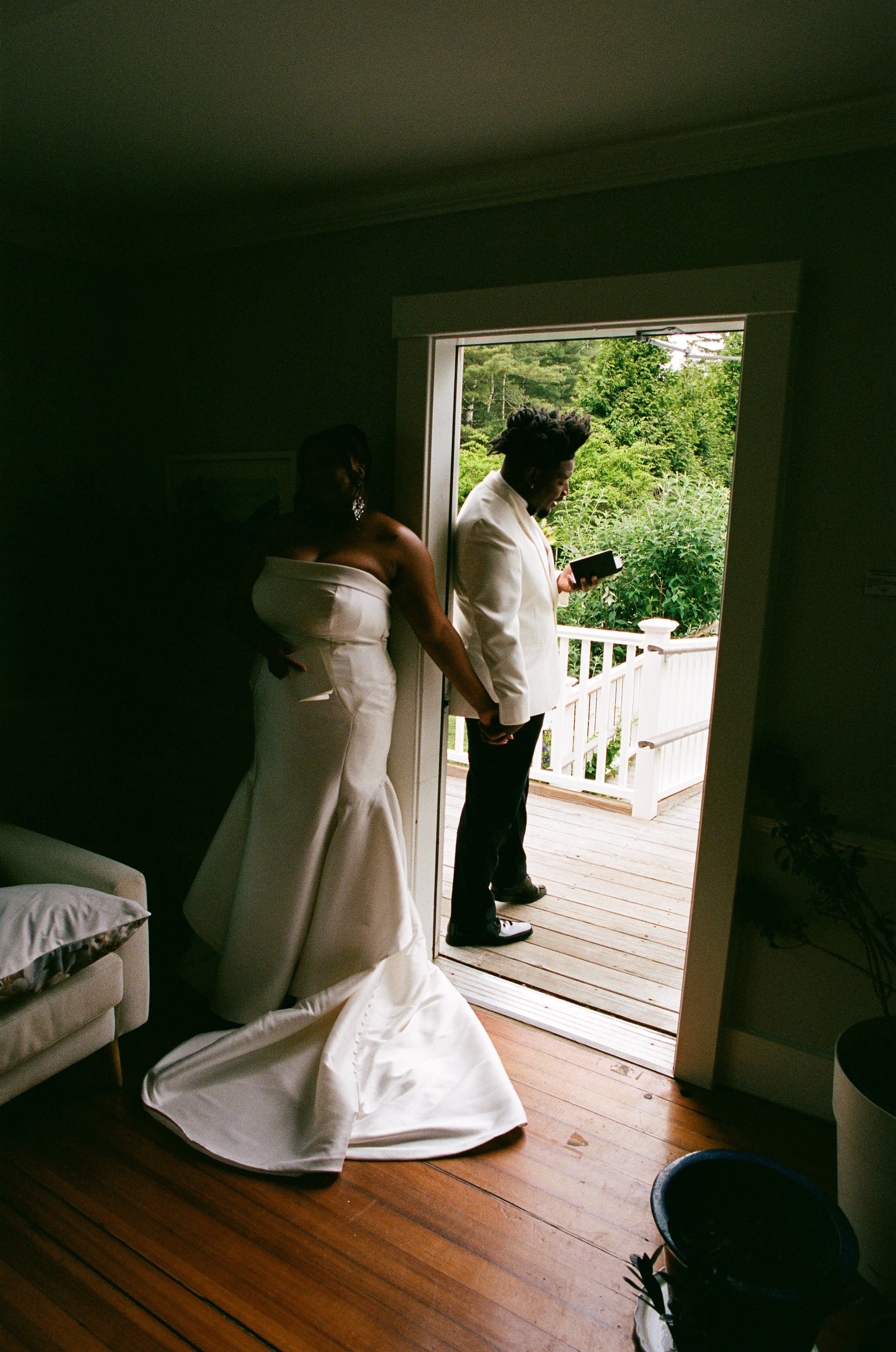 A woman in a white dress standing inside a dimly lit room and a man in a white suit and black shoes standing outside on a wooden balcony, both holding hands. The woman appears to be looking down, and the man is looking at his phone. The room opens to