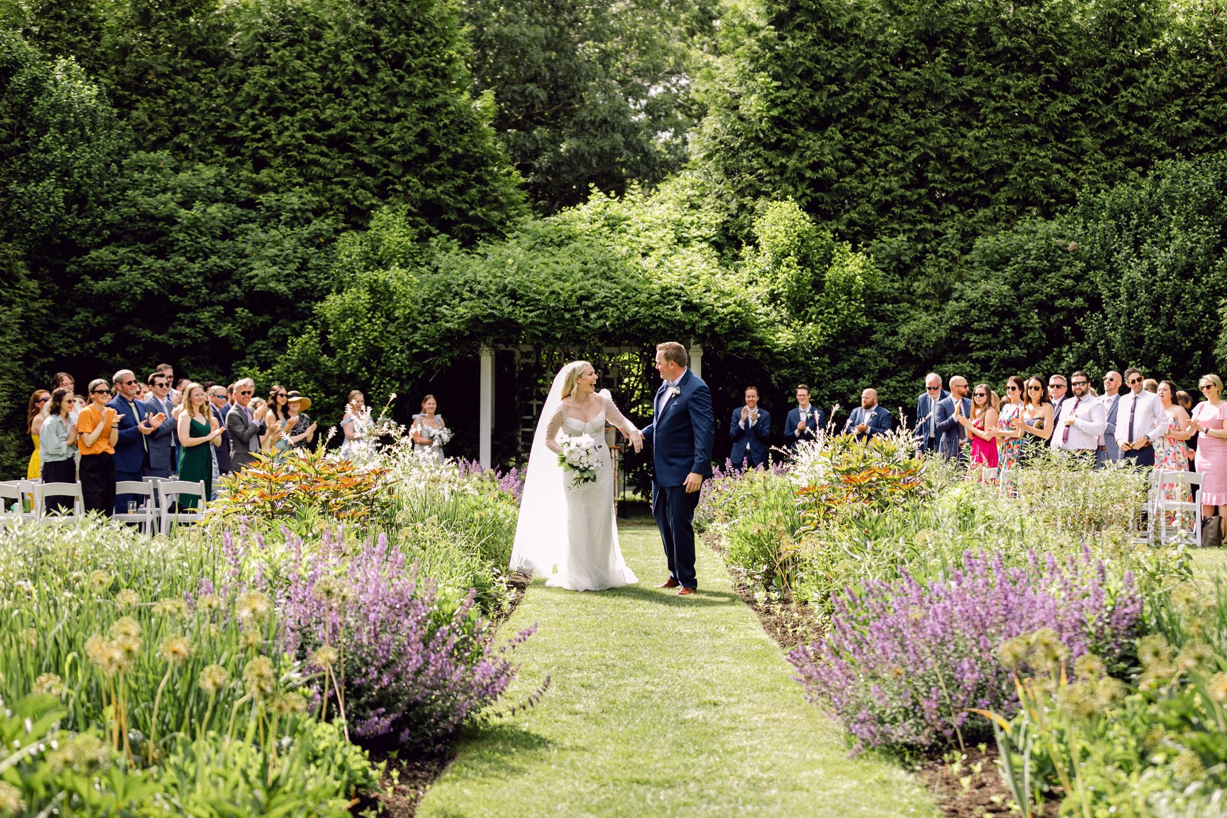 A bride and groom holding hands during their wedding ceremony outdoors at Lambert's Cove Inn on Martha's Vineyard, surrounded by guests and lush green trees, with purple and white flowers lining the aisle.