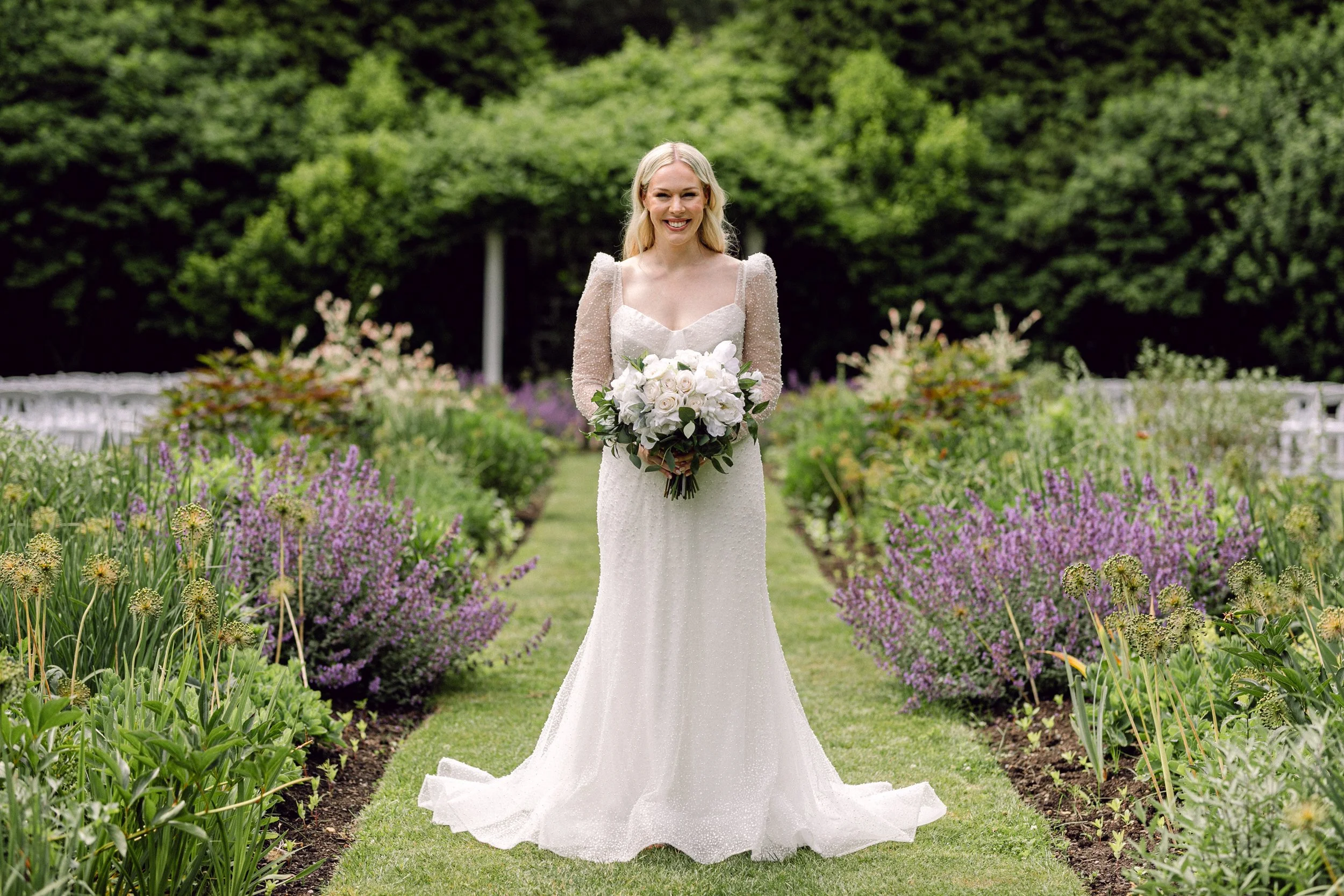 A smiling bride in a white wedding dress holding a bouquet of white flowers in a garden with purple and green plants at Lambert's Cove Inn on Martha's Vineyard.