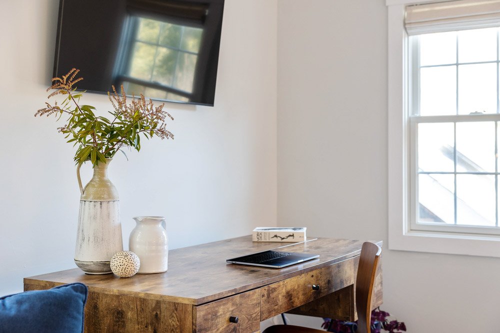 A wooden desk with a laptop, decorative books, and white vases with green foliage, in a room with a window and a wall-mounted TV.