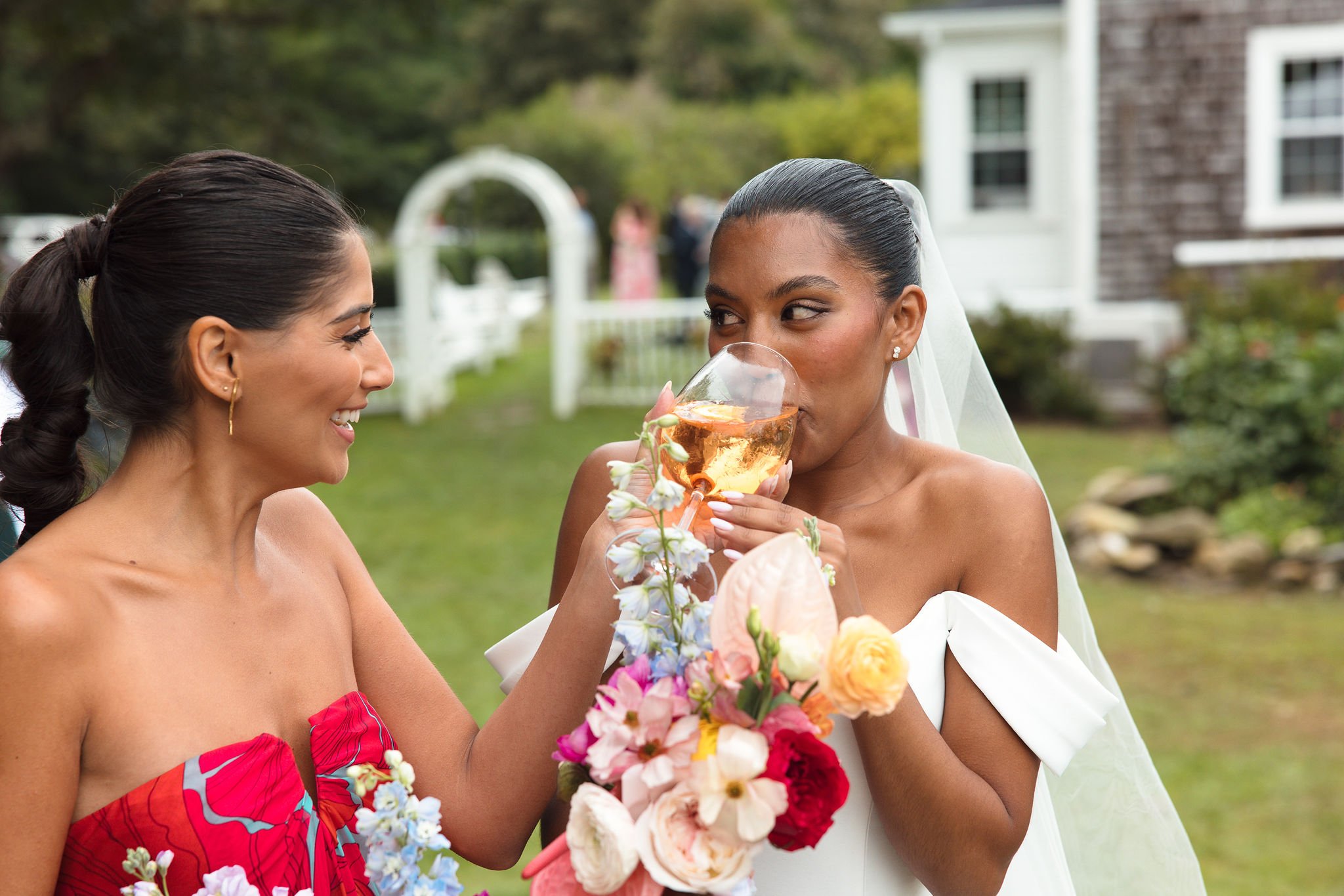Two women with dark hair wearing dresses, one in red and the other in white, at an outdoor wedding reception. The woman in white is holding a champagne glass with a floral arrangement, and they are smiling and looking at each other.