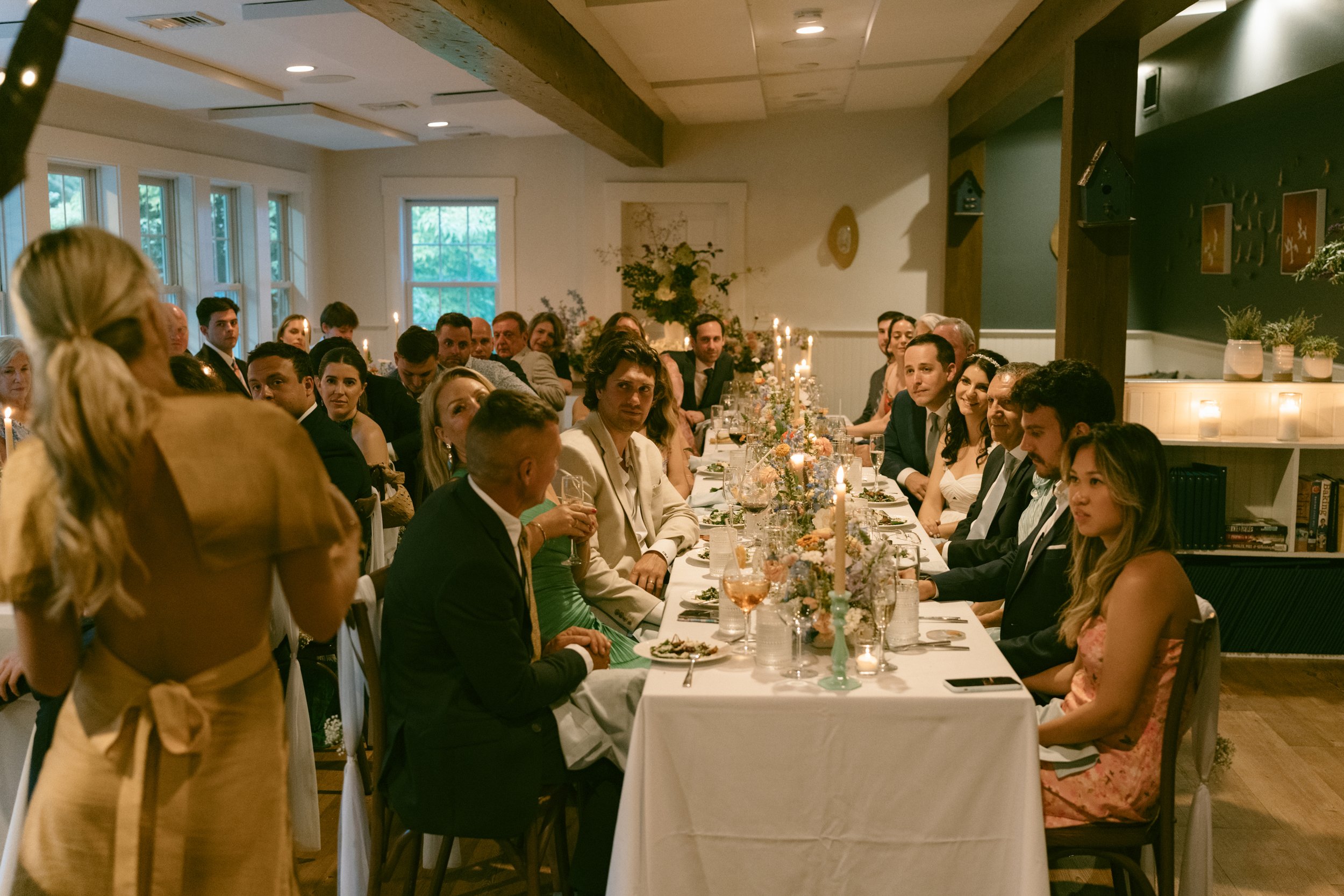 A large group of people dressed formally sitting at a long dining table during a celebration or event in a well-lit room with large windows and floral decorations at Lambert's Cove Inn on Martha's Vineyard.