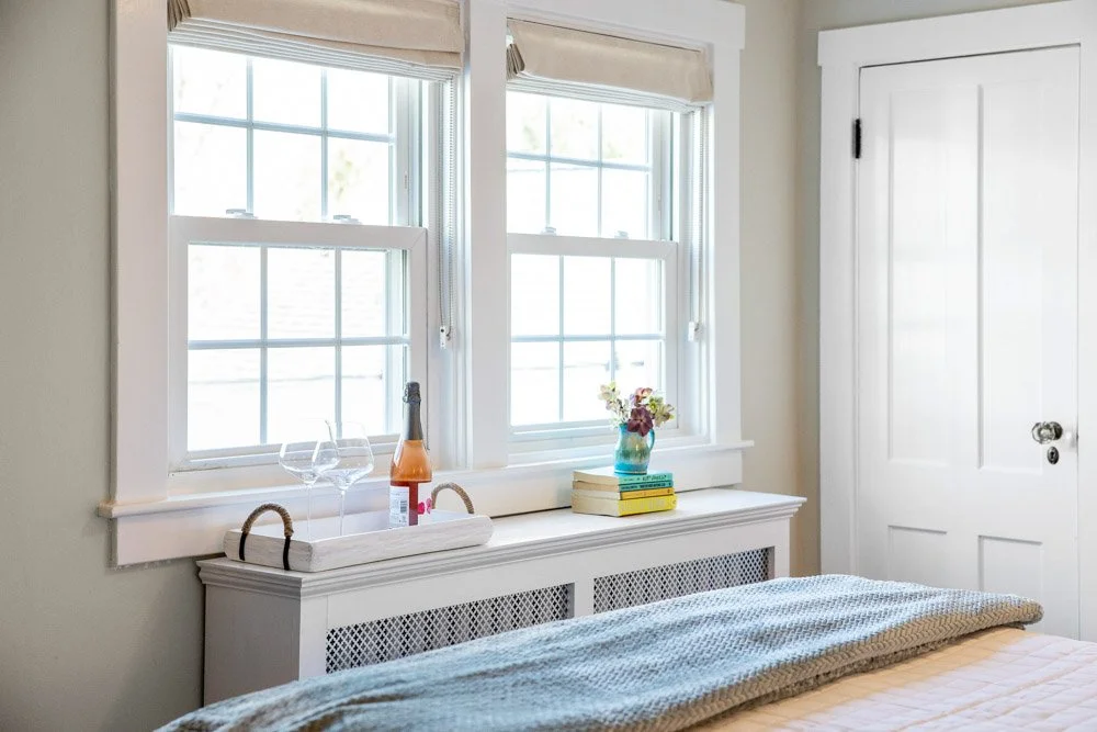 A bedroom with two large windows, a white radiator cover with lattice panels, a bed with a textured blanket, a tray with a bottle of champagne and two glasses, a stack of books with a vase of flowers, and a closed white door.