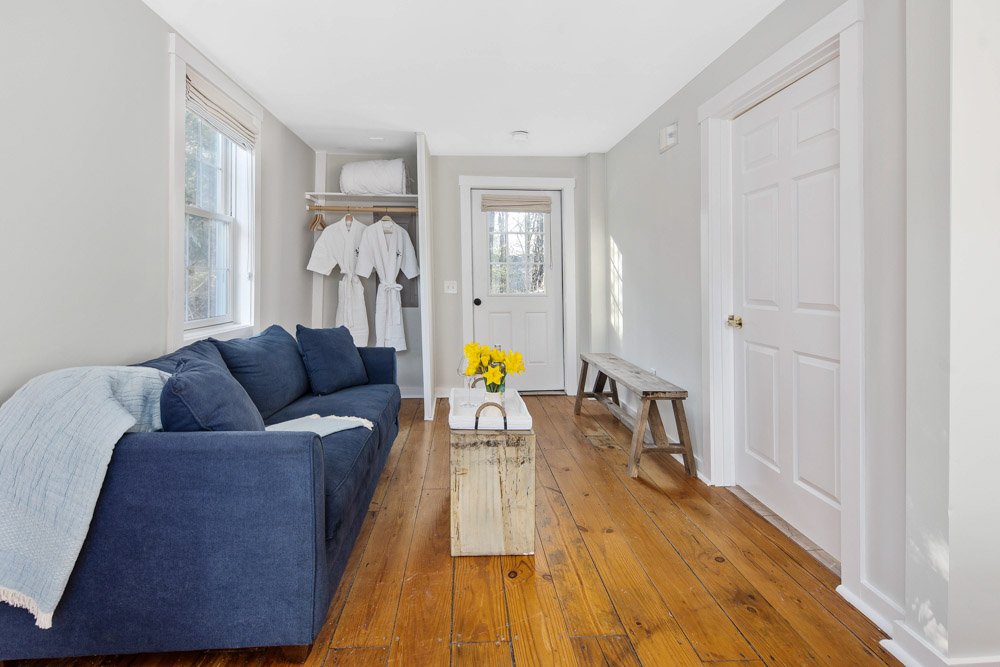 A cozy living room with a navy blue sofa, wooden floor, white walls, a small wooden bench, a white door, and a small shelf with two white robes hanging and a rolled towel on top. There is a window with blinds and a vase of yellow flowers on a wooden 