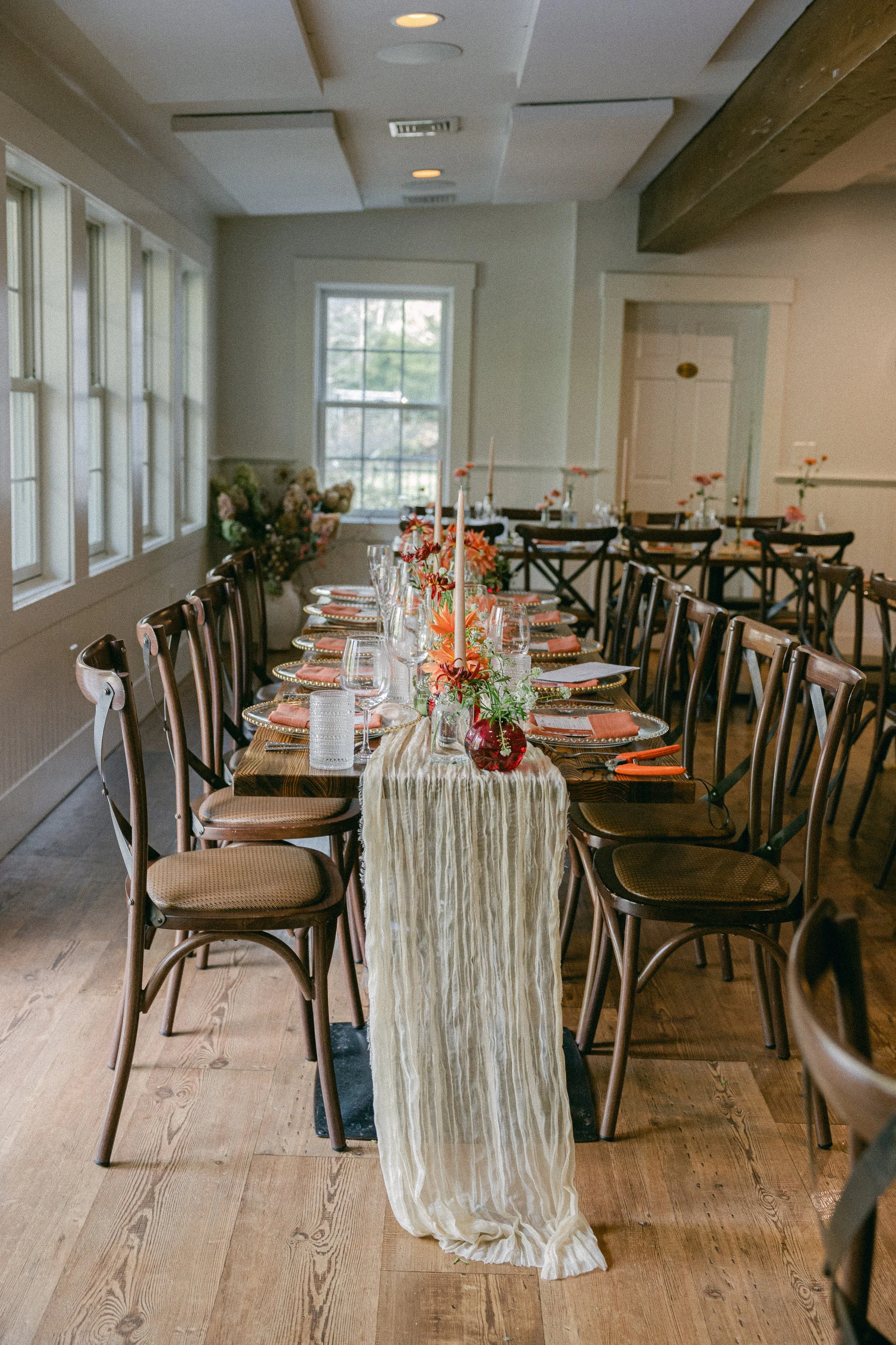 A long wooden dining table set for a formal event with orange flowers in glass jars, candles, and place settings including glasses and napkins. The room has wooden floors, large windows, and a white textured table runner hanging down the center of th