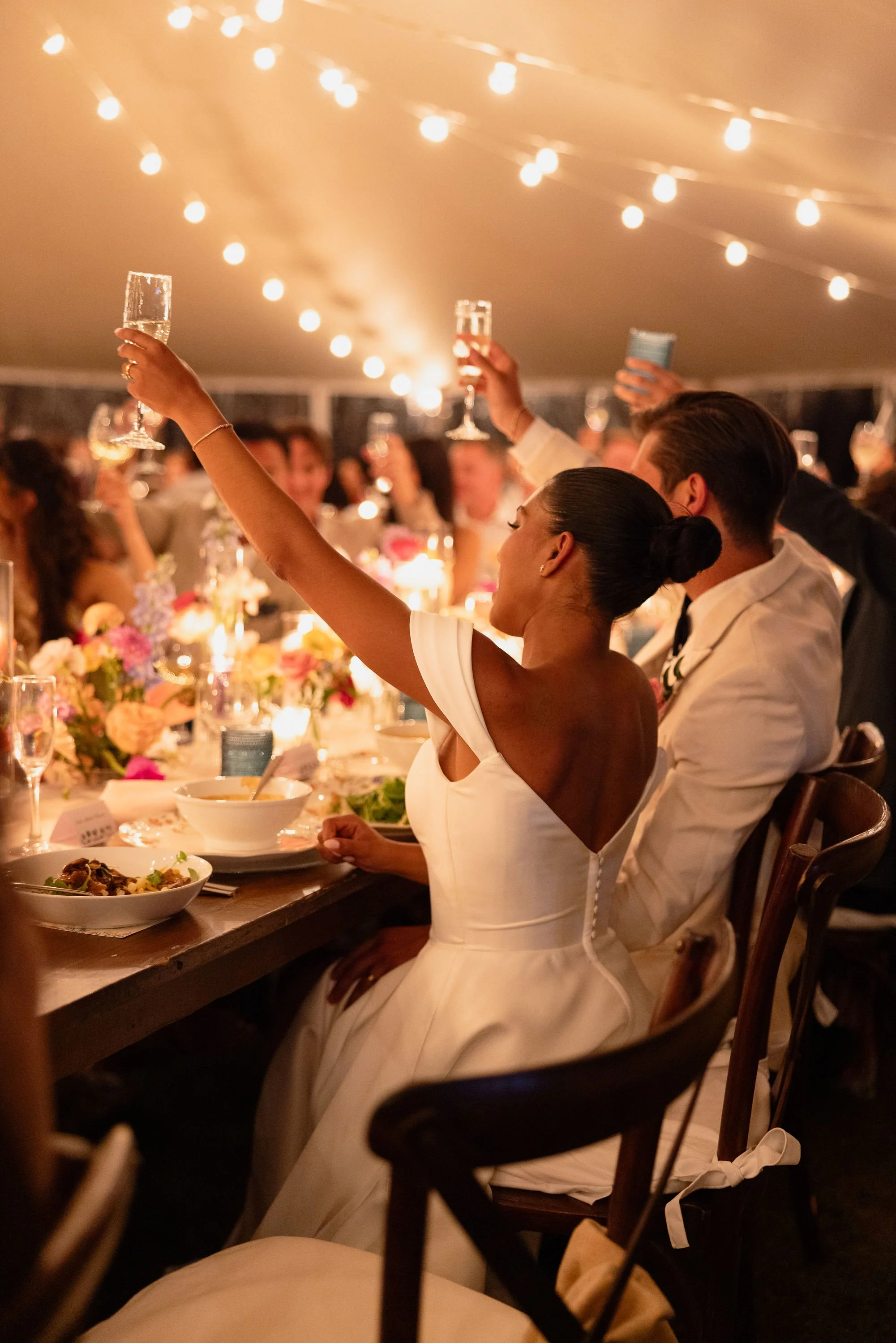 A woman in a white dress raising a champagne glass at a celebration dinner with string lights overhead, surrounded by other people holding drinks at Lambert's Cove Inn on Martha's Vineyard.