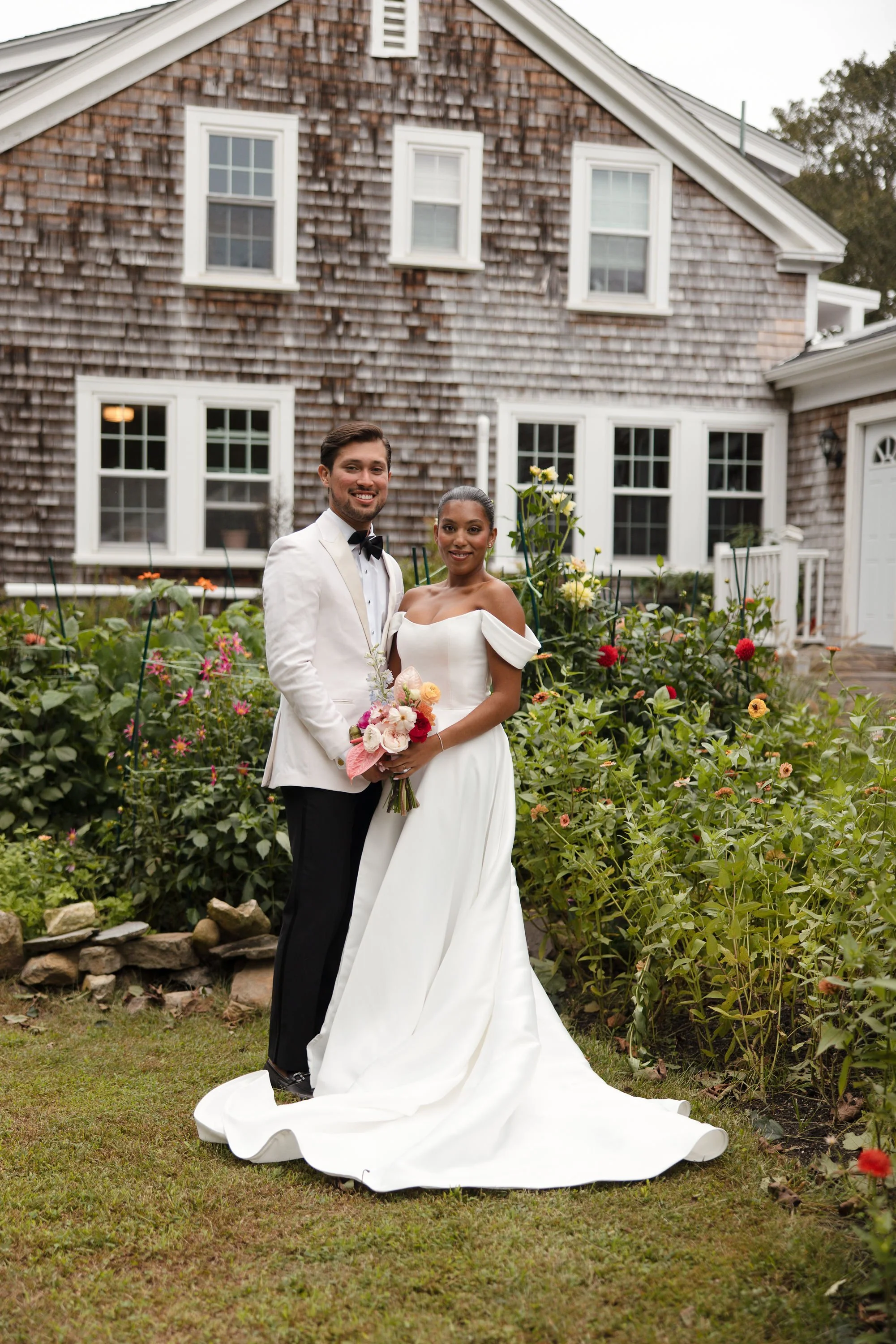 A newlywed couple standing in a garden in front of a wooden house, holding a bouquet of flowers, smiling at the camera during a wedding at Lambert's Cove Inn on Martha's Vineyard.