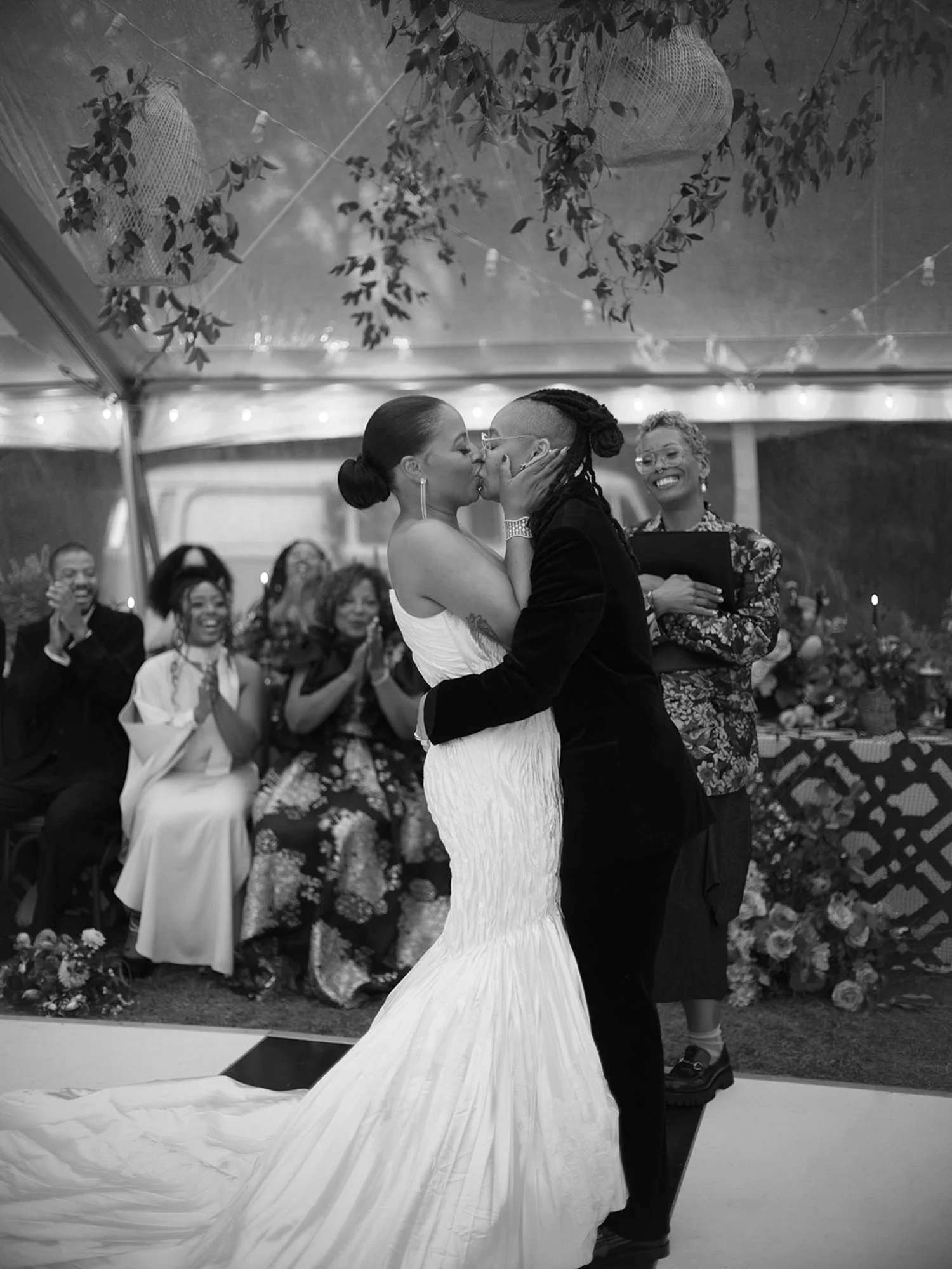 A couple shares a kiss at their wedding ceremony at Lambert's Cove Inn on Martha's Vineyard, with friends and family applauding in a decorated outdoor setting.