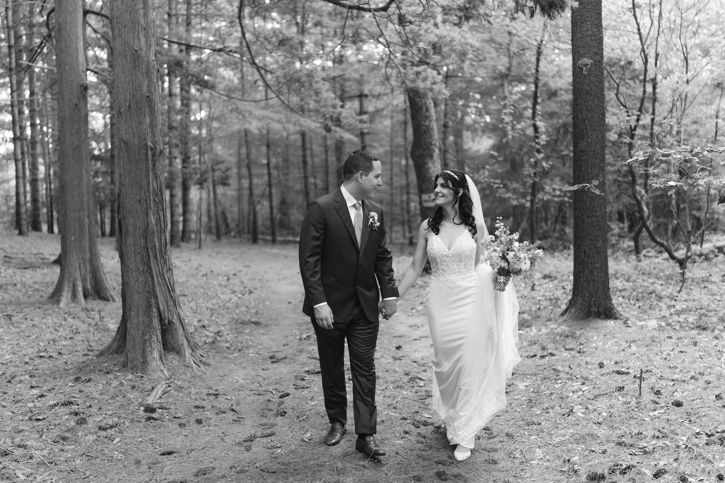 A black-and-white photo of a couple in wedding attire walking hand in hand through a woods at Lambert's Cove Inn on Martha's Vineyard. The bride is wearing a strapless wedding dress and holding a bouquet, while the groom is dressed in a suit.