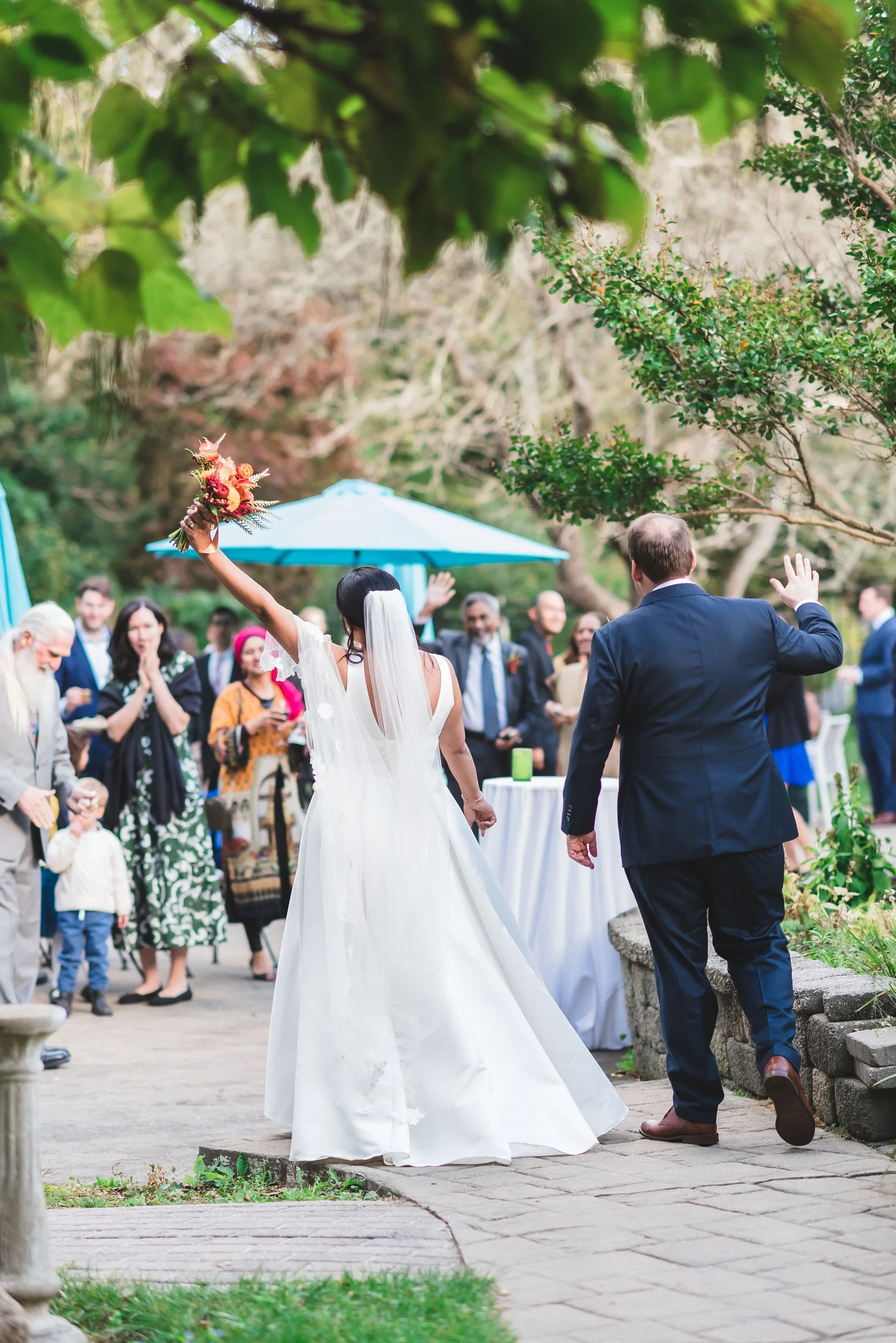 A bride in a white wedding dress holding a bouquet raises her arm, while a groom in a navy suit walks beside her with his hand raised. Guests are gathered in the background outdoors during daytime, with some clapping and taking photos.