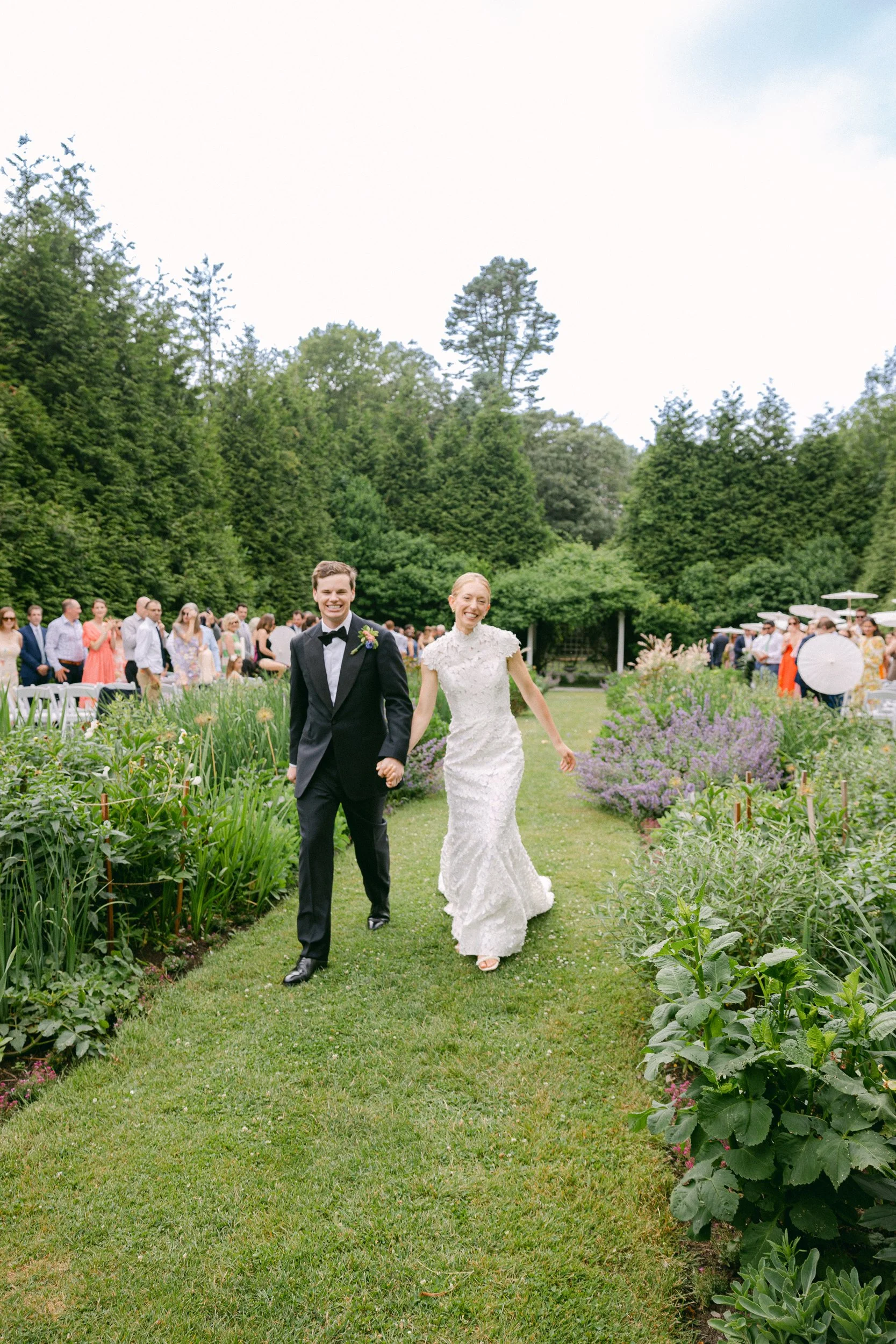 A bride and groom walking hand in hand on a lush green garden path during a wedding at Lambert's Cove Inn on Martha's Vineyard, with guests in the background, some holding umbrellas, and tall trees surrounding the area.